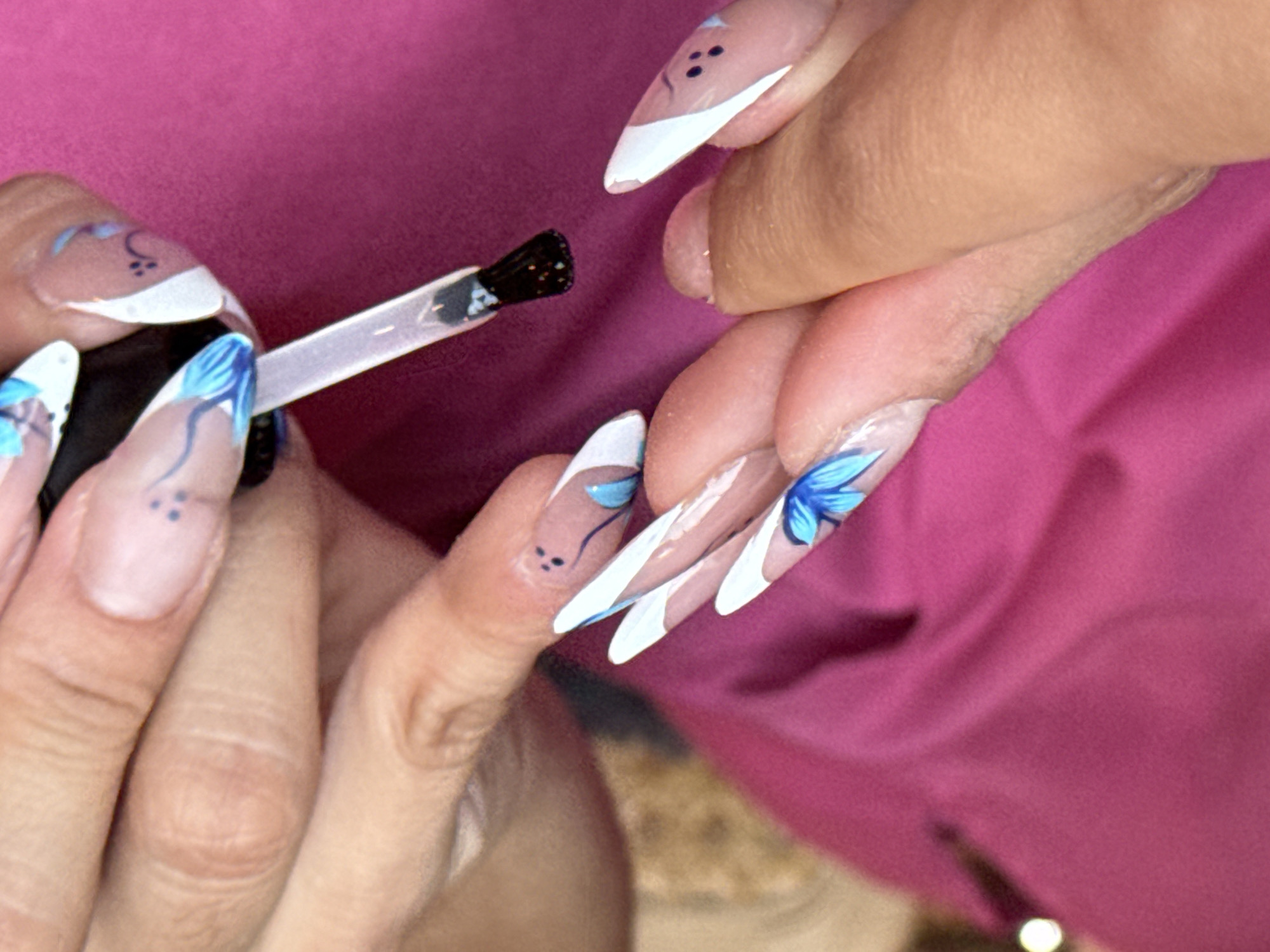 A close-up shot of a person applying a top coat to acrylic nails. The nails are pointed and have a white tip design with blue flower details. The person applying the top coat is holding a brush with a clear, shiny liquid on it. The background is a blurred pink fabric. The location in the city of Puebla, Mexico, is not discernible from this image.