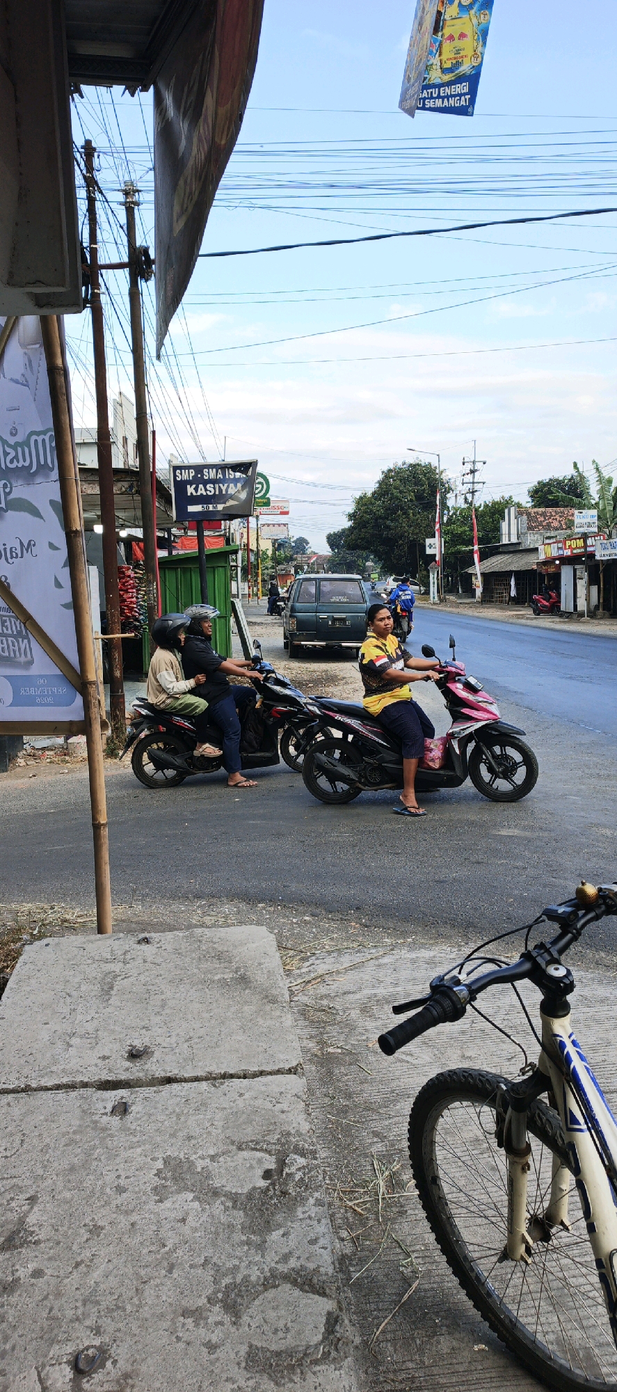 The image depicts a street scene in Kasiyan, Indonesia. In the foreground, a bicycle is partially visible, leaning on its side. Behind the bicycle, a person on a pink scooter, wearing a yellow and black shirt and dark shorts, is riding down the road. To the left of the scooter, another scooter carries two people, one driving and one passenger, both wearing helmets. Further down the road, a dark green SUV is parked, and in the background, there are various shops and buildings lining the street. A signpost indicating "SMP - SMA ISLAM KASIYAN" and "50 M" is also visible. The sky is clear with a few wispy clouds, and power lines crisscross the scene. The overall impression is that of a typical day in a small Indonesian town.