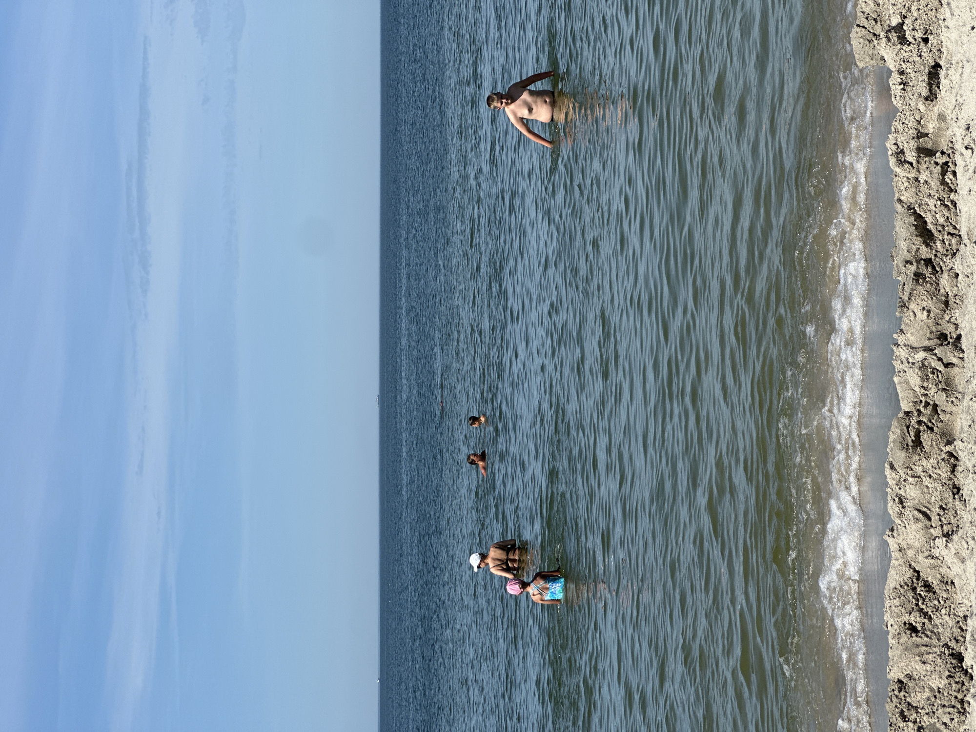 The image depicts a sandy beach with people wading in the calm water. The scene is set during the daytime, under a clear sky, suggesting pleasant weather. The water is a deep blue, with gentle ripples on the surface. The sand is light brown and textured, with small dunes visible along the edge of the water.
Several individuals are scattered across the water, appearing to be enjoying a swim or simply cooling off. One man, standing waist-deep, is facing away from the camera and seems to be interacting with someone in the water. Further out, other people are visible as smaller figures in the distance. Closer to the shore, a woman wearing a pink swim cap and a patterned swimsuit is standing in the water, facing the camera. Another person, wearing a white cap, is partially submerged next to her.
The overall impression is one of a relaxed summer day at the beach, with people enjoying recreational activities in a natural setting. The location is identified as Baltiysk, Russia. There is no visible text in the image.