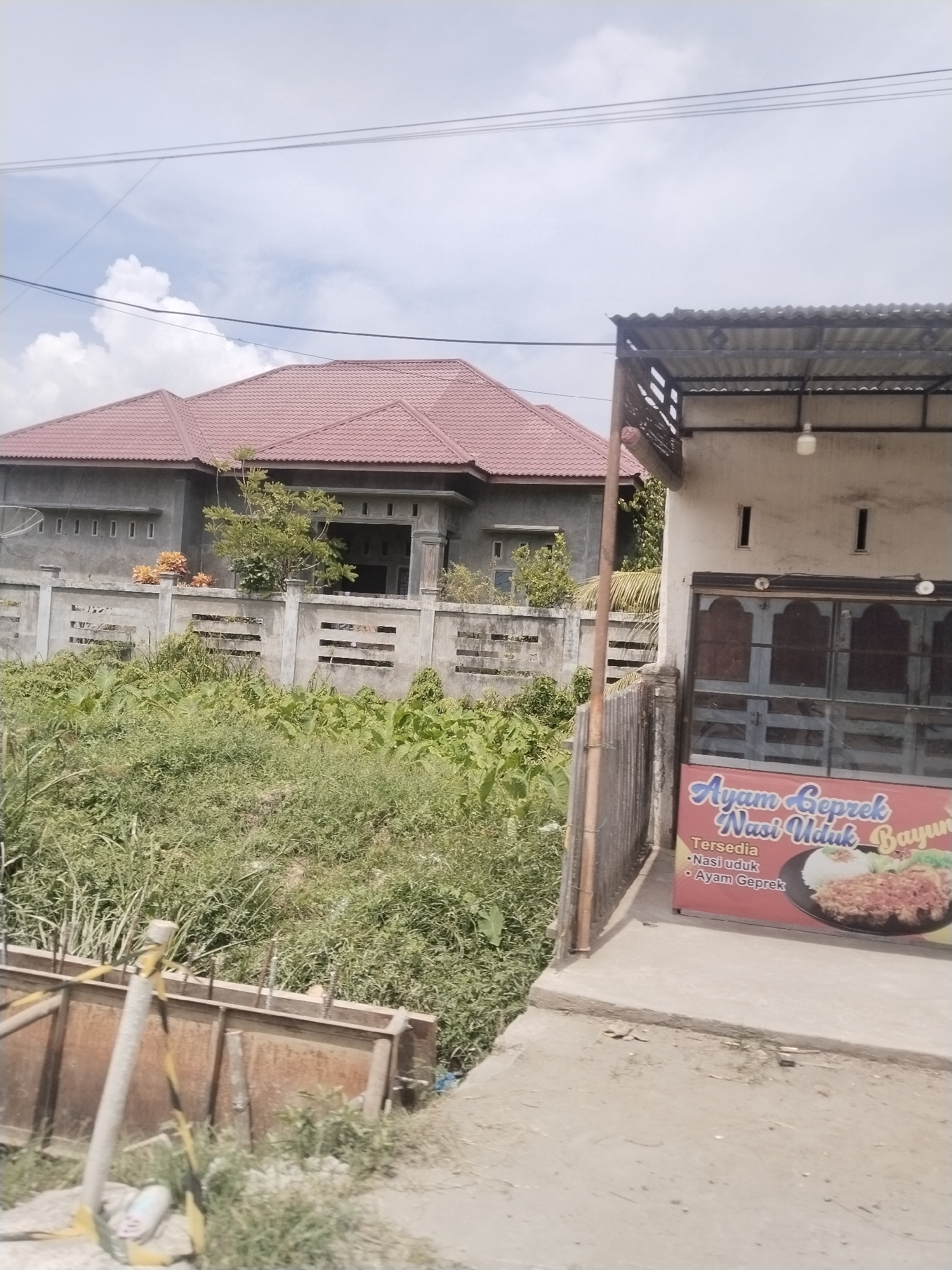 A sunlit outdoor scene depicts a residential building with a red tiled roof in the background, flanked by a concrete fence. In the foreground, a building with a corrugated metal roof overhang houses a food stall. A banner on the stall advertises "Ayam Geprek Nasi Uduk Bayu," indicating the availability of fried chicken with coconut rice and a signature dish. The surrounding environment is overgrown with lush green vegetation. The image appears to be taken in the city of Beurandang, Indonesia, suggesting a street view of a local eatery and its surroundings.