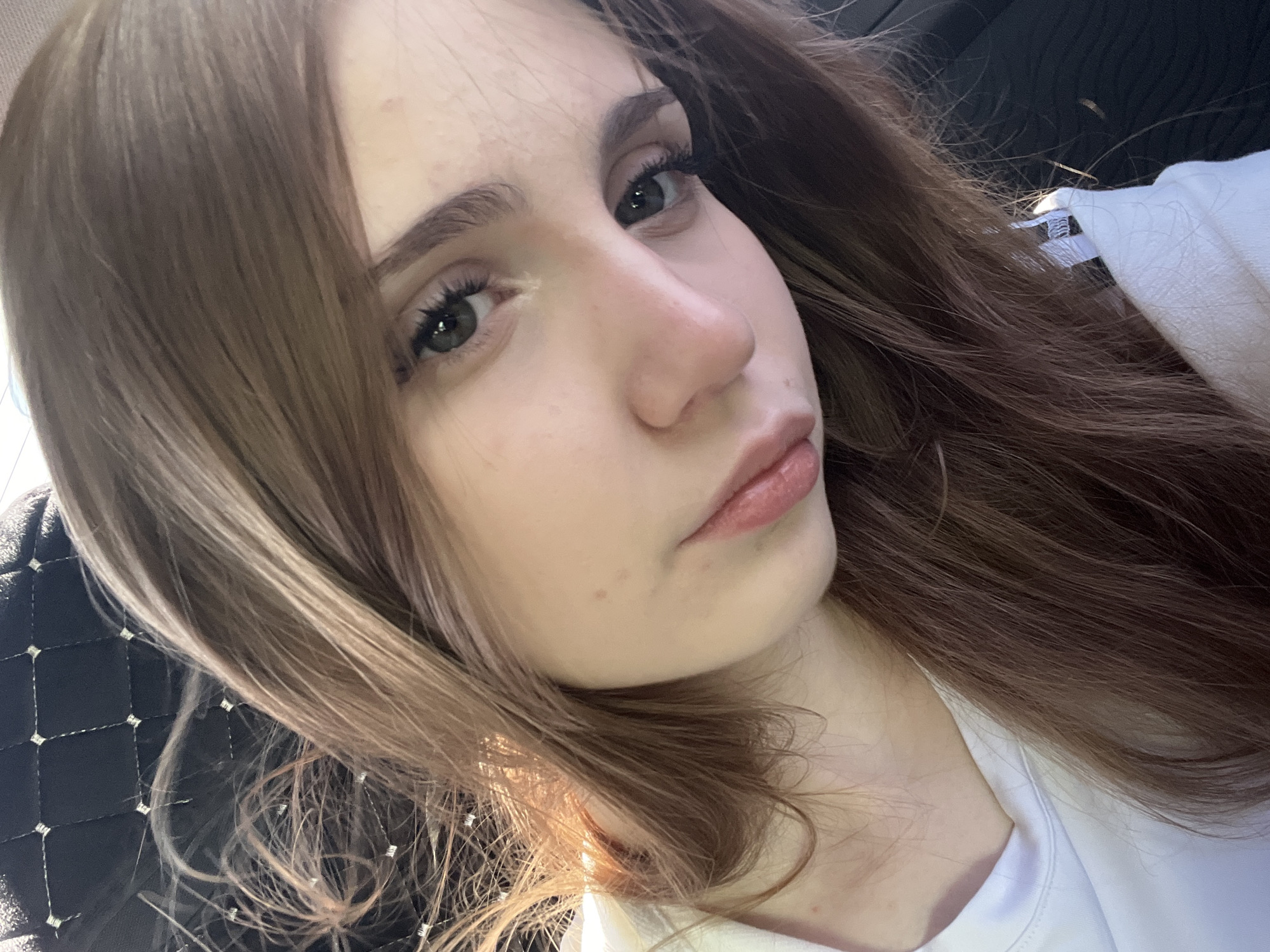 The image is a close-up, selfie-style shot of a young woman's face. She has long, light brown hair that is slightly windswept. Her eyes are green, and she is wearing light makeup, including mascara. Her lips are slightly parted and appear to have a glossy finish. She is wearing a white t-shirt. The background is dark and out of focus, with a textured pattern that suggests the interior of a car, possibly a headrest. There is no discernible information in the image that would place it specifically in the city of Orenburg, Russia.