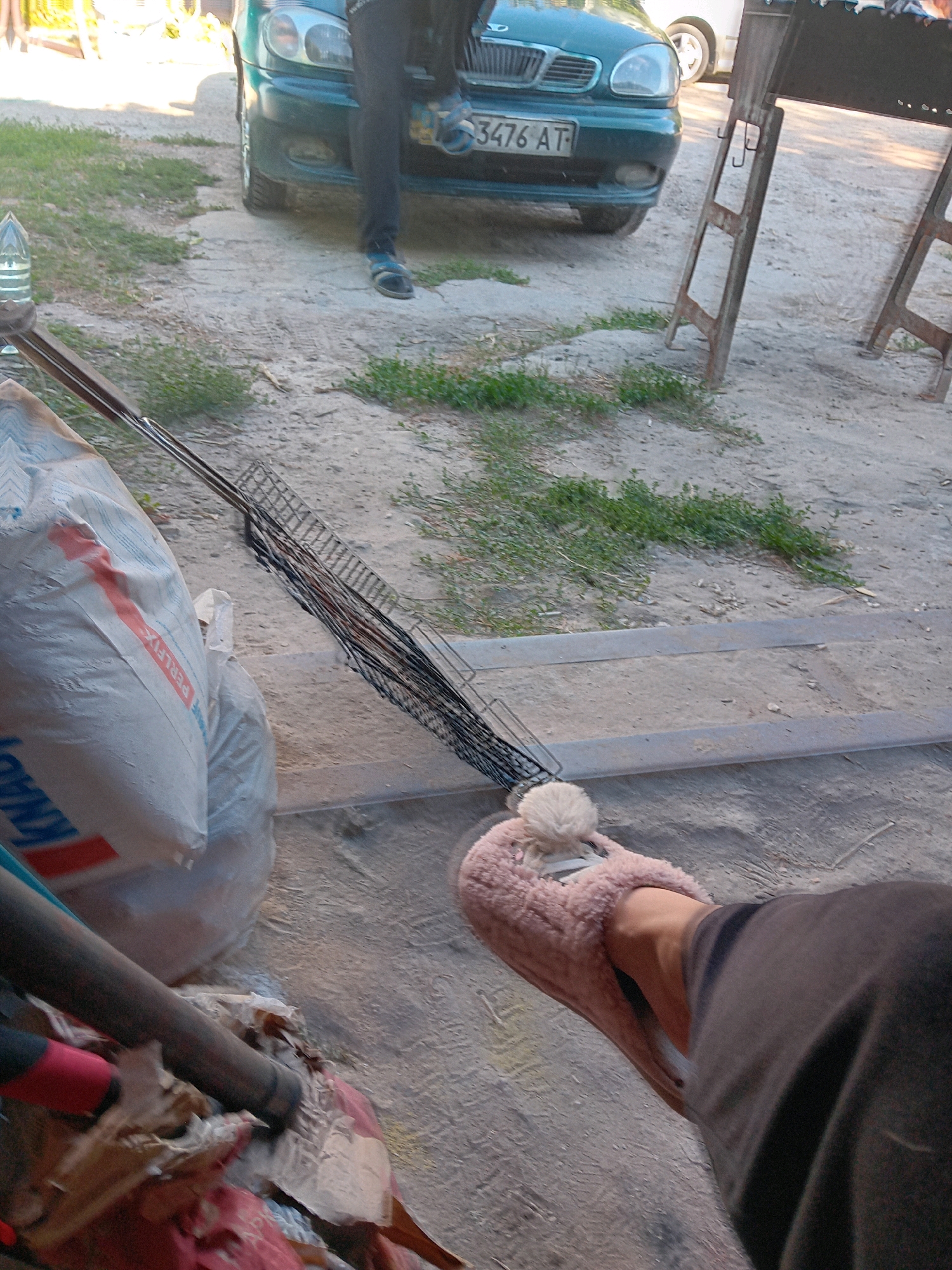 The image depicts an outdoor scene with a green car in the background. A person's legs and feet are visible in the foreground. The person is wearing dark pants and a fuzzy pink slipper with a pom-pom. To the left of the person's feet is a large white bag, possibly containing building materials, with red and blue lettering. A metal grate is positioned behind the bag, extending out of frame. The ground is a mixture of dirt and gravel with patches of green grass. The scene appears to be in a residential or utilitarian area, possibly a yard or a street in Chuhuiv, Ukraine, given the context. The lighting suggests it is daytime, with shadows indicating the sun's position. There is no apparent interaction between the visible person and any other individuals in the scene.