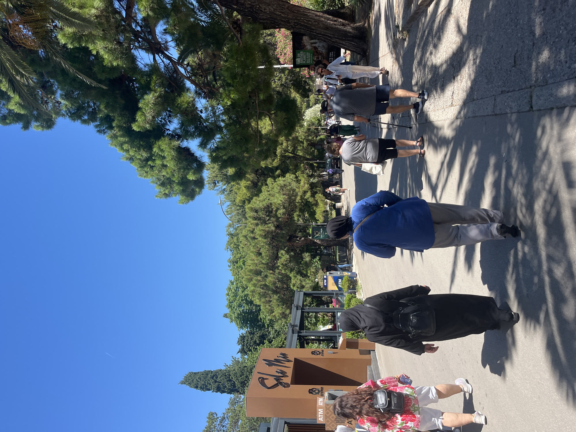 This is a full, eye-level, outdoor shot of a group of people walking on a paved walkway in Budva, Montenegro. The image is taken on a sunny day, as indicated by the bright blue sky and the shadows cast by the trees and people.
In the foreground, three people are walking away from the camera. The person on the left wears a brightly colored floral print shirt and white shorts, with a black backpack. The person in the middle is wearing a long black abaya and a small black backpack. The person on the right wears a blue shirt and gray pants.
Further down the walkway, several other individuals are visible, some walking and some standing. To the left of the walkway, there is a building with the name "Sole Mio" prominently displayed. ATMs are visible on the side of this building. Tall green trees line the walkway, casting dappled shadows on the ground. On the right side of the walkway, a large tree with dense foliage dominates the scene. A billboard with text and images is visible behind this tree. In the distance, more trees and buildings can be seen. The overall scene depicts a public area with pedestrian activity and commercial establishments.