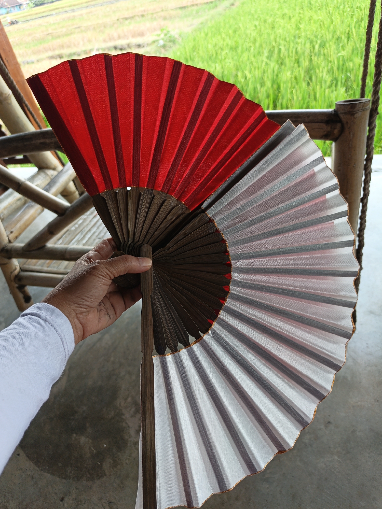 In Sekarsuli, Indonesia, a hand wearing a white long-sleeved shirt holds a two-toned folding fan outdoors. The fan is split vertically, with the left side featuring vibrant red fabric and the right side a translucent white fabric. Both sides are pleated and attached to a dark wooden frame. The fan is positioned in front of a blurred background of green rice fields and a bamboo structure, suggesting a rural or semi-rural setting. The ground beneath the fan appears to be concrete or dirt. The lighting suggests it is daytime.