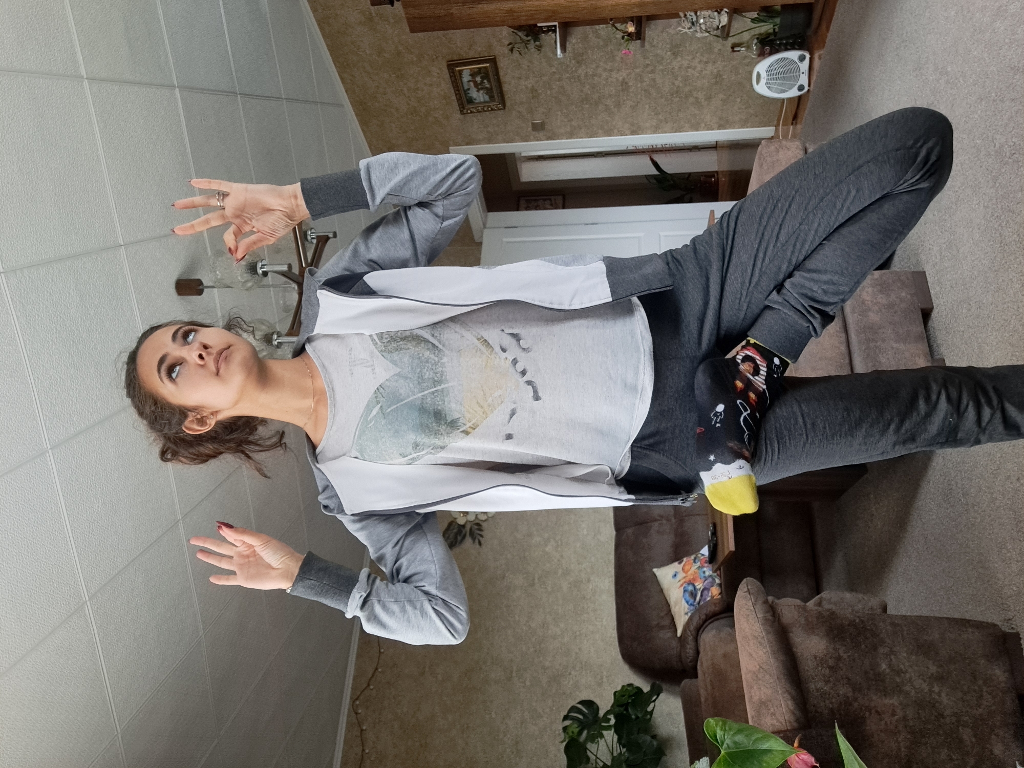 A woman is indoors, standing near a ceiling with square tiles, and a lit candelabra. She is wearing gray sweatpants, gray sleeves, and a white jacket. She has a ring on her right hand. She is wearing a sock with images printed on the side with a yellow toe. A brown couch is behind her, as well as a plant, table, and a small fan. The room has tan carpeting. There is a brown cabinet in the background with a framed picture on the wall. There are additional potted plants on the top of the cabinet. The location is unknown; there is no evidence to support that this scene takes place in Negovan, Bulgaria.