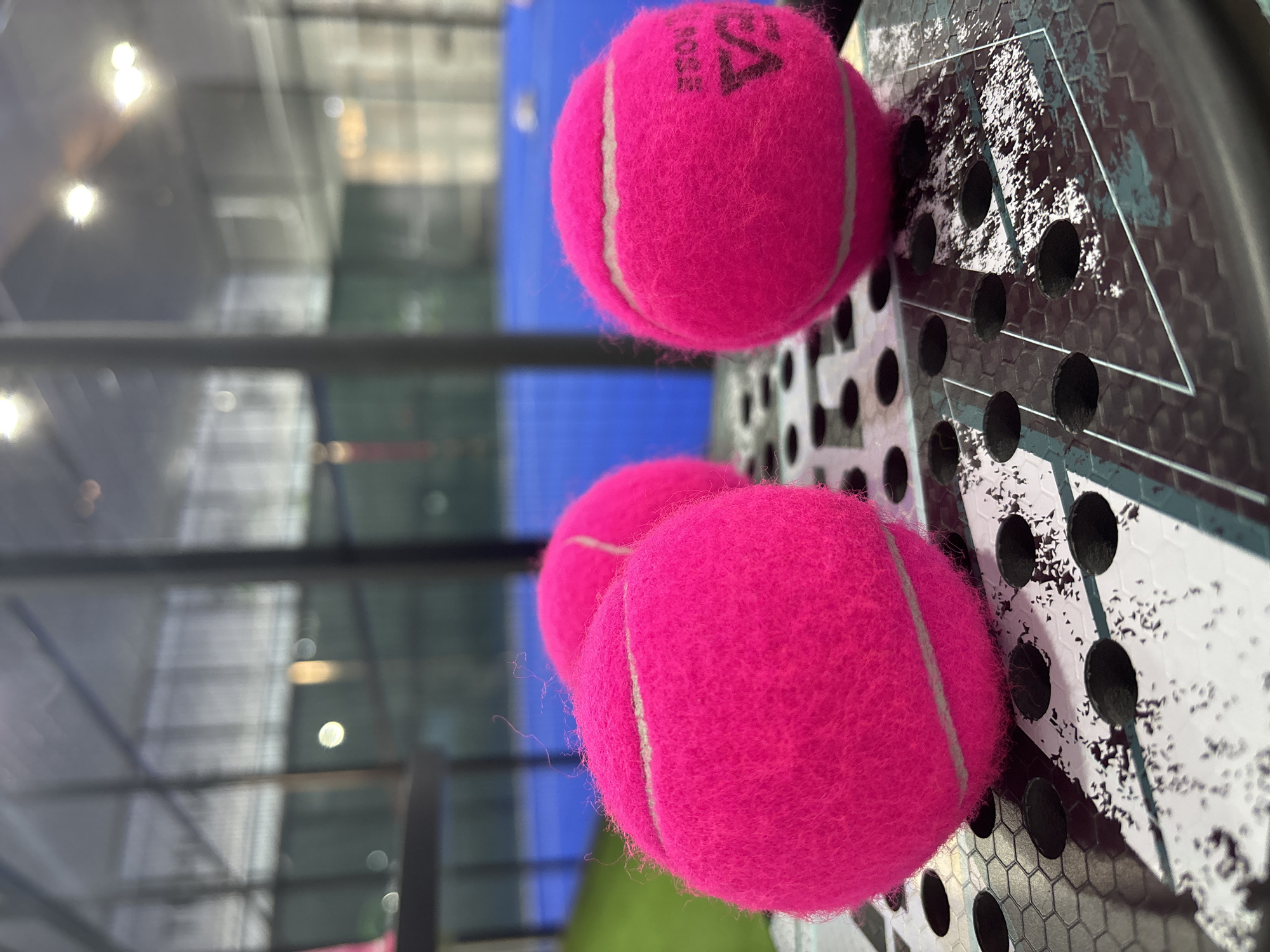 Three bright pink tennis balls are positioned on the surface of a black padel racket. The racket, with its distinctive hexagonal pattern and a graphic design, rests on what appears to be a padel court. In the background, a blue court surface is visible, bordered by green turf. The overall scene is set indoors, within a facility that likely houses multiple courts, suggested by the presence of large glass windows and overhead lighting. The lighting in the background is diffused, indicating artificial illumination within the enclosed space. The image does not contain any individuals or show any ongoing activity; it is a static composition of sporting equipment within a sports venue.