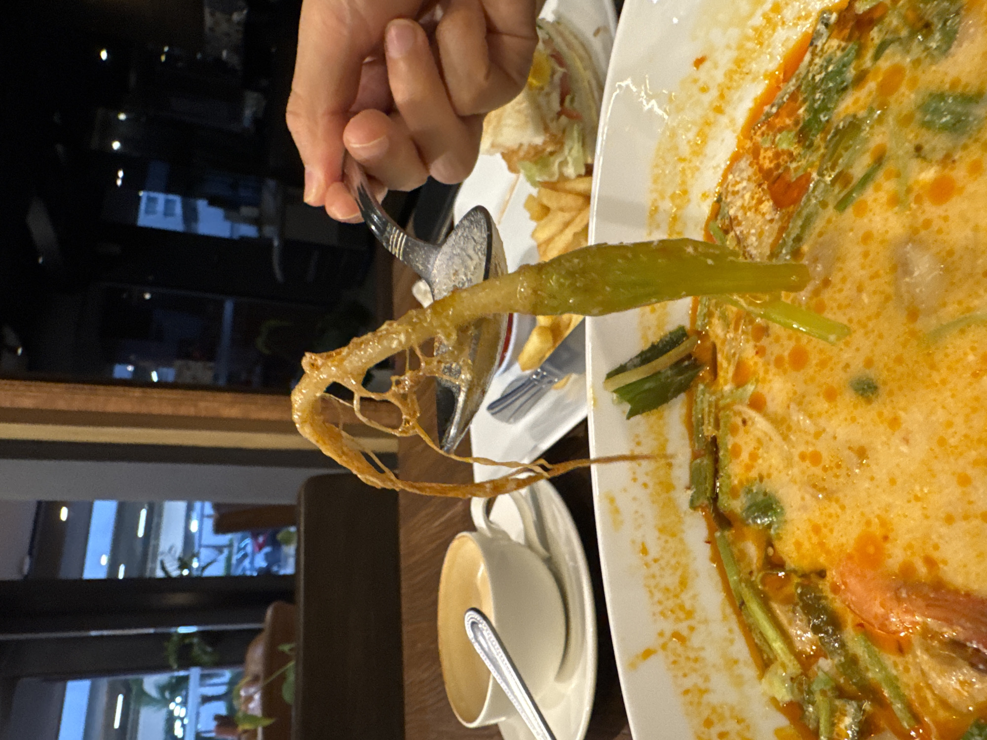 A person's hand holds a spoon over a bowl of orange-colored soup with green vegetables, lifting a piece of food that resembles a crispy, golden-brown deep-fried element with tendrils. In the background, a white teacup with a latte sits on a saucer, and a plate with french fries and a sandwich is visible. The environment appears to be a restaurant with wooden tables and chairs, and large windows showing a blurred outdoor scene with streetlights. The overall impression is a casual dining setting, potentially in Bangkok, Thailand, given the presence of what appears to be a Thai dish.