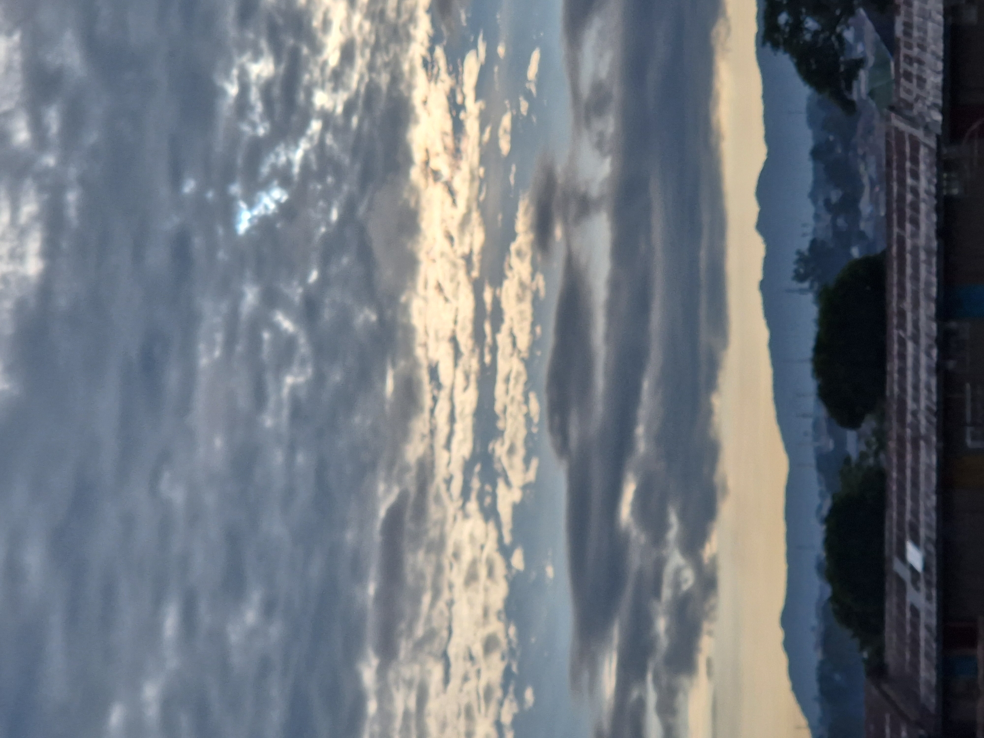 The image depicts a blurry, overcast sky above a distant mountain range. The sky is filled with numerous clouds of varying densities, with some areas appearing brighter where sunlight is filtering through. Below the sky, a dark, silhouetted mountain range stretches across the horizon. In the foreground, the tops of buildings with visible roofs are present, suggesting an urban or suburban setting. The overall impression is of a calm, slightly somber, early morning or late afternoon scene. The location is specified as Unknown, Unknown.