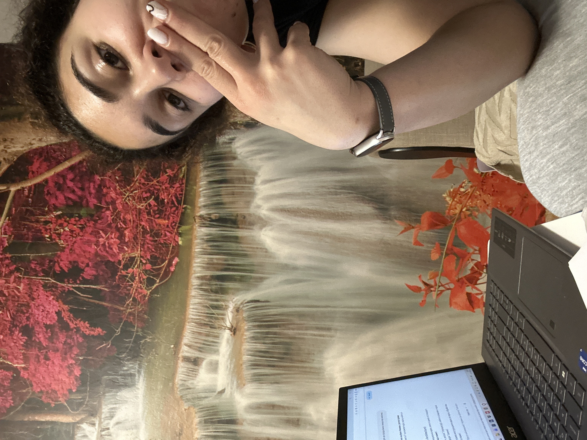 Here is a concise description of the image:
The image shows a woman from a high angle, with her face partially obscured by her hand, wearing a smartwatch on her left wrist. She appears to be working on a laptop. The laptop is on a table or surface featuring a landscape image with red leaves and what seems to be a waterfall. A vase with artificial orange flowers is also near the laptop.