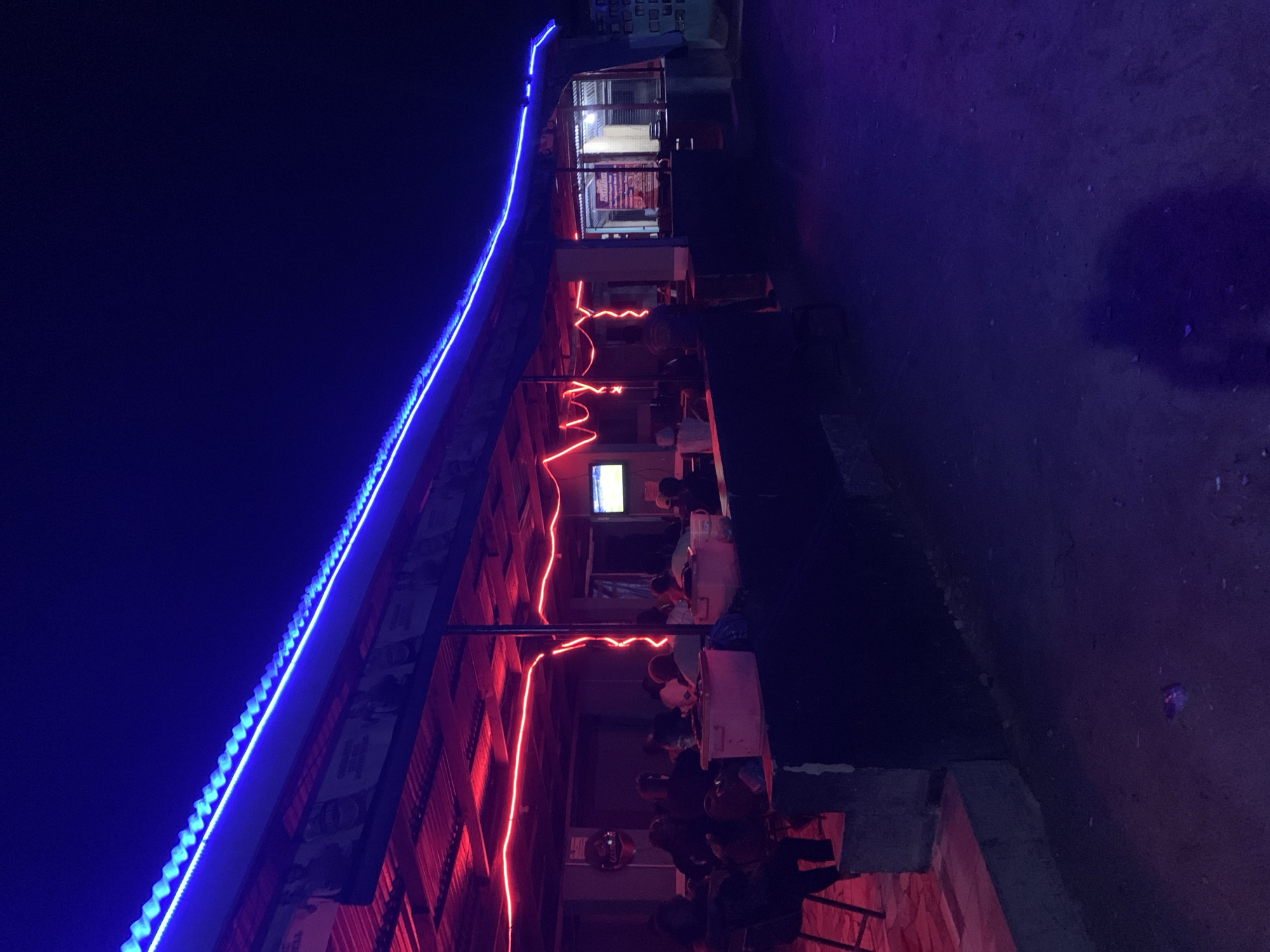 The image shows an outdoor eatery in New Stadium, Nigeria at night. The building has a corrugated metal roof outlined with blue LED lights. Red neon lights vertically illuminate the seating area. Under the roof, people are seated at tables, some watching a television mounted on the wall. The ground appears to be paved. The overall lighting is dim with a mix of blue, red, and the dim light from the interior of the building.