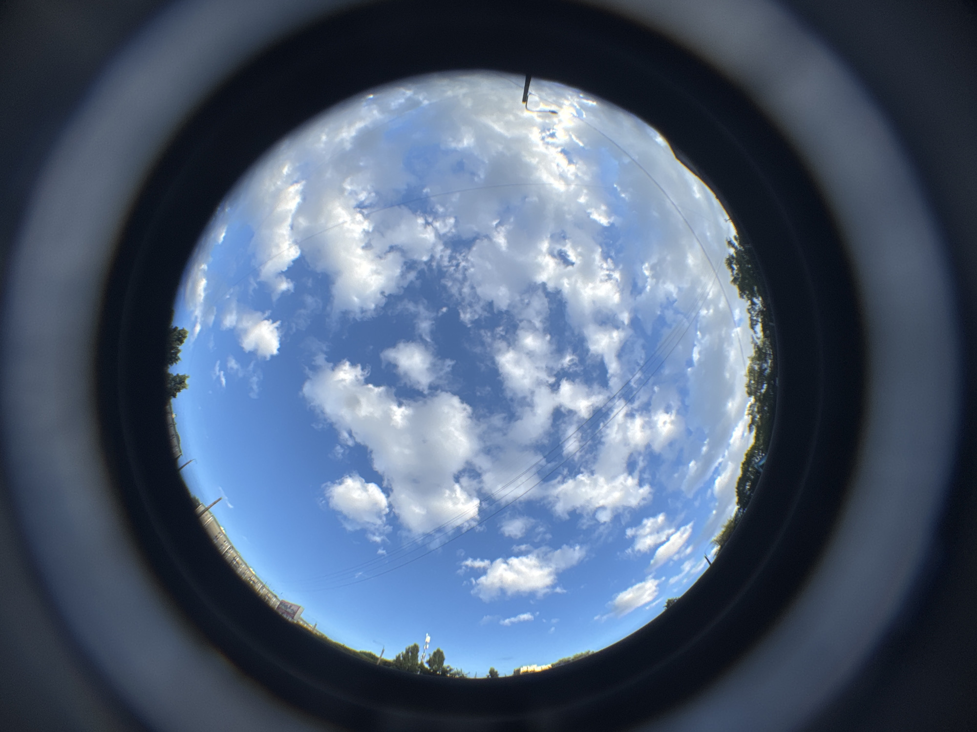 The image captures a distorted, fisheye perspective of a bright blue sky filled with scattered white clouds. In the lower portion of the frame, a dark, circular border encloses the sky, suggesting the image was taken through a lens or opening. A thin line of trees is visible along the horizon at the very bottom of the sky. Three parallel power lines stretch across the sky, and in the upper left quadrant, what appear to be buildings or structures are faintly discernible. On the right side, a thin, dark object extends into the sky from the edge of the frame, possibly a piece of equipment or a shadow. The overall impression is of an outdoor scene under a partly cloudy sky, with the context of Saint Petersburg, Russia, though no specific landmarks are clearly identifiable.