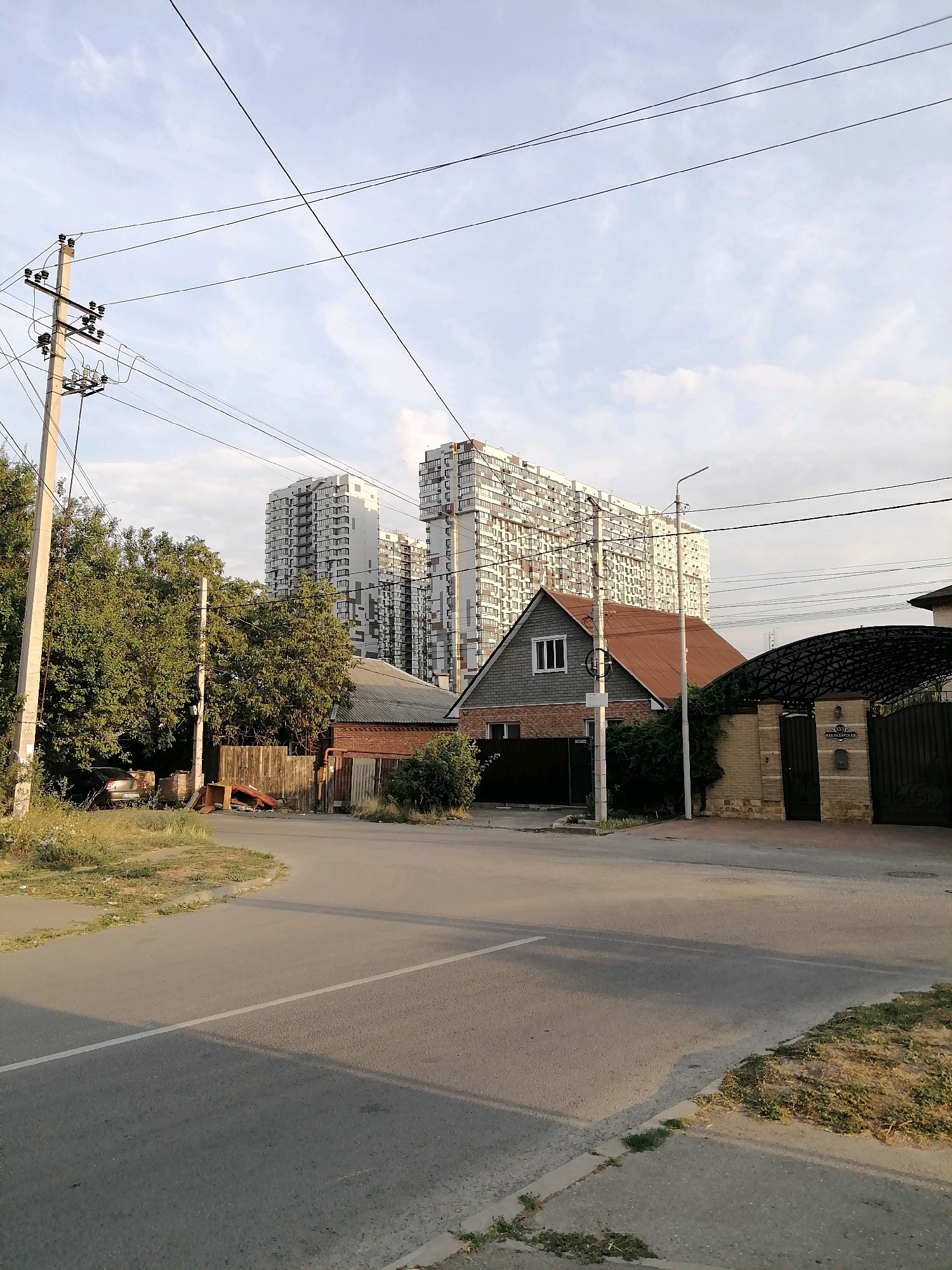 The image shows a street scene in Rostov-on-Don, Russia, with a focus on a residential area. In the background, several tall apartment buildings with white facades and numerous windows dominate the skyline. In the foreground, a paved road curves to the right. On the left side of the road, there is a concrete utility pole with electrical wires stretching across the frame. Adjacent to the pole is a dense cluster of trees, with a silver car parked among them. A wooden fence and some discarded materials are visible behind the trees.
To the right of the road, a single-family house with a reddish-brown pitched roof and gray siding is visible. It has a large window on the upper level and a smaller window on the lower level. Next to this house, a brick wall encloses a property with a dark metal arched gate. Above the gate, there is a sign with a street number and street name, which appears to read "ул. Ленина 389/1". A street light is also positioned near the gate. The overall environment suggests a mixed residential area with both new high-rise developments and older single-family homes. The sky is partly cloudy, indicating daytime.