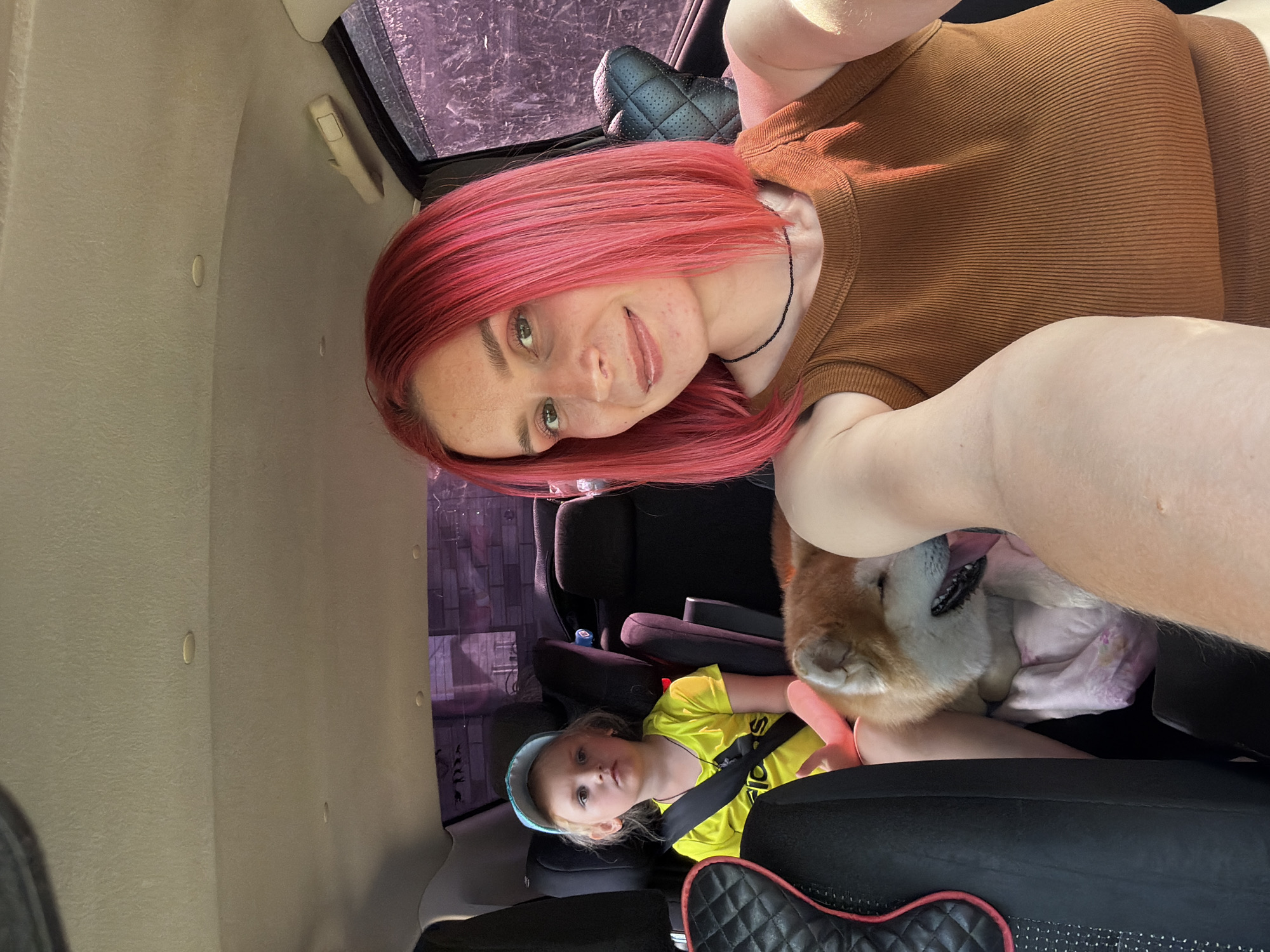 The image depicts a selfie taken inside a vehicle, likely a car, with a young child and a dog visible in the background. The primary subject is a woman with bright pink hair, smiling at the camera. She is wearing a brown, ribbed, sleeveless top and a black necklace. Her arm is extended to capture the selfie.
In the background, a young girl with blonde hair and a light blue headband is seated in a car seat, wearing a bright yellow t-shirt with black writing on it. A dog, appearing to be a Shiba Inu with its characteristic reddish-brown and white fur, is nestled next to the woman, with its head resting on her arm and tongue slightly out. The interior of the car is visible, with car seats and parts of the vehicle's interior trim. The background outside the window suggests an outdoor setting with trees or foliage, and the lighting indicates it is daytime.
The overall mood appears to be casual and familial, perhaps during a road trip or outing. The subjects seem relaxed and comfortable. The image is taken in Chaikovsky, Russia, as indicated by the provided location context. The visible text on the girl's shirt appears to be "adidas".