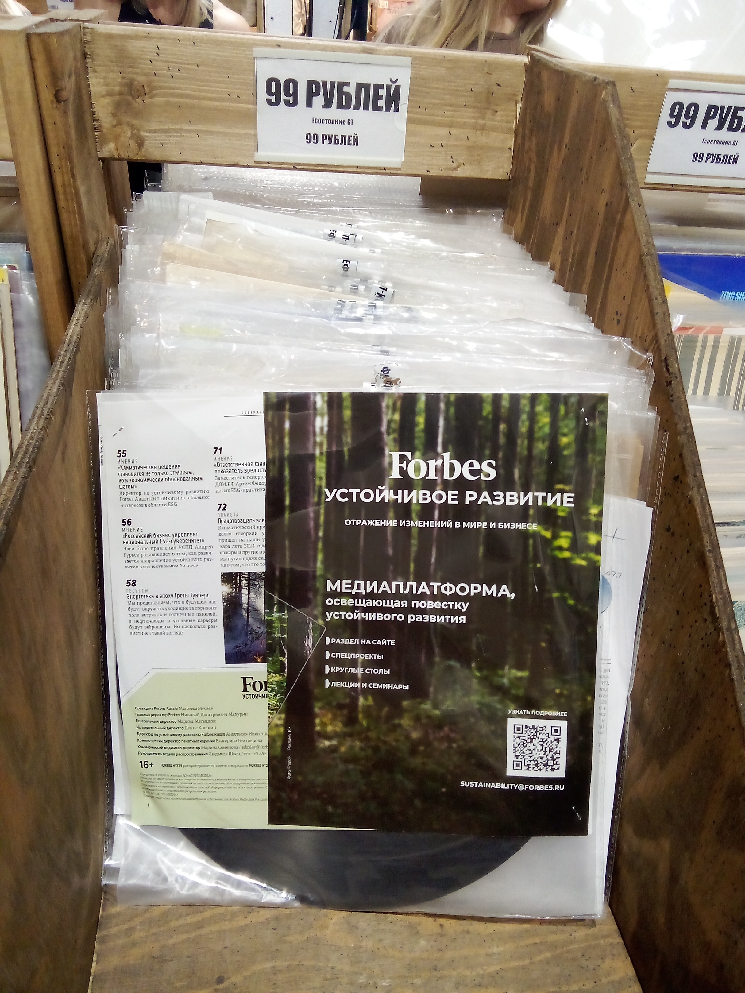The image shows a close-up, eye-level, indoor, daylight shot of a wooden display rack filled with magazines. The magazines are protected by plastic sleeves. On the top shelf of the display, there are two price tags indicating "99 РУБЛЕЙ" (99 Rubles). One price tag is on the left side of the display, and another is on the right. The magazines in the display are oriented vertically, with their covers facing outwards. The dominant magazine in the foreground has a dark green background with an image of a forest and text in Russian. The text on this magazine includes "Forbes" and "УСТОЙЧИВОЕ РАЗВИТИЕ" (Sustainable Development). Below this, there is a QR code and the email address "SUSTAINABILITY@FORBES.RU". The overall environment appears to be a retail setting, possibly a bookstore or newsstand.