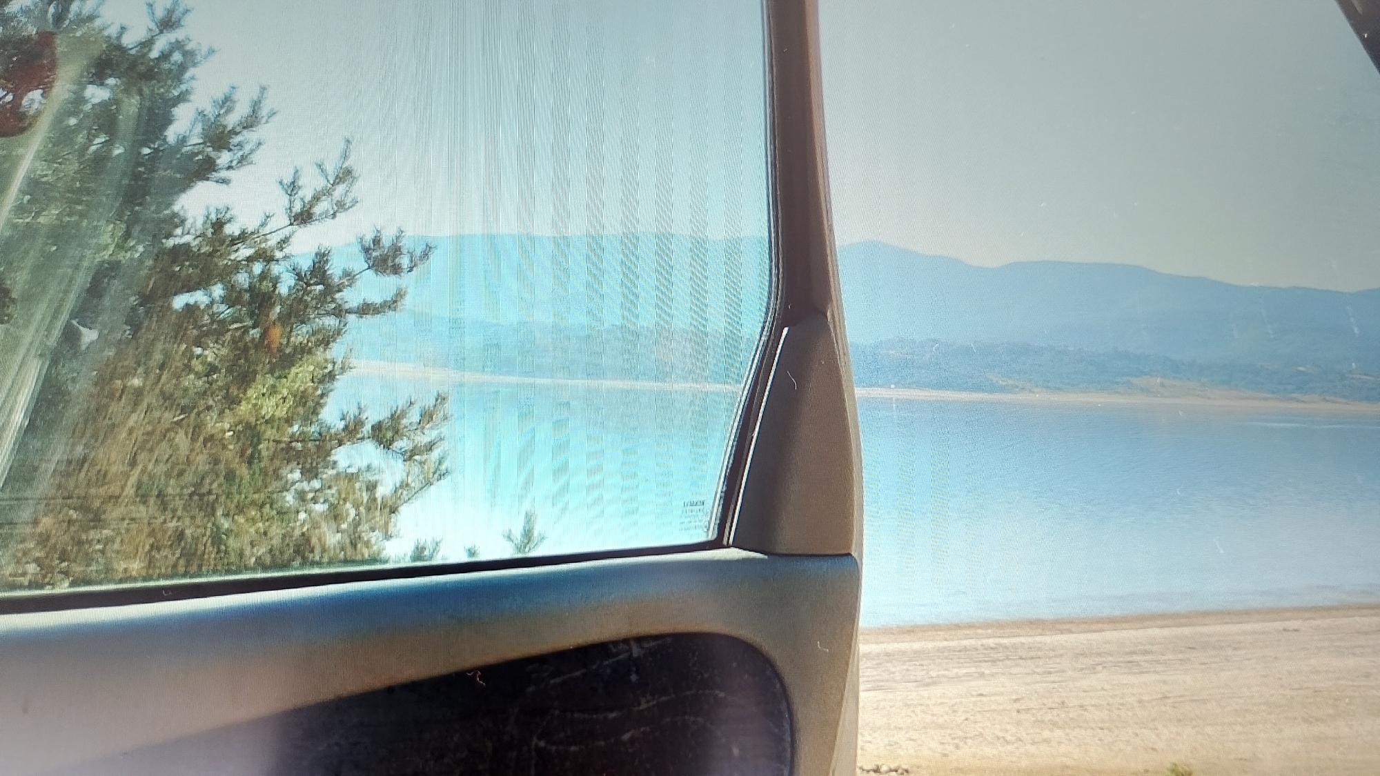 The image captures a view through a car window, looking out at a landscape. The foreground shows the interior of a vehicle, with the armrest and door panel visible in the lower portion. The car window reflects light and shows a slight wavy pattern, suggesting it might be a digital screen or a slightly distorted window.
Through the window, a body of water, likely a lake or reservoir, is visible. The water is a vibrant turquoise color. On the far side of the water, there are hills or mountains covered in lush green vegetation. The sky is a hazy light blue, indicating a possibly overcast or distant view. On the left side of the frame, a portion of a tree with green foliage is visible, seemingly close to the car. The overall scene suggests an outdoor setting, possibly a scenic overlook or a stop along a route near a body of water and natural landscape.
The location is specified as Smolyan, Bulgaria. Smolyan is known for its picturesque mountainous terrain, deep valleys, and numerous lakes and reservoirs, such as the Dospat Reservoir, Yundola Reservoir, and Shiroka Polyana Reservoir. The vegetation visible in the image is consistent with the flora found in the Rhodope Mountains, which surround Smolyan. The presence of a car and the perspective from a window are typical of a travel or sightseeing scenario. The hazy distant mountains are characteristic of the Bulgarian landscape, particularly in the Rhodope region, which often experiences atmospheric conditions that create a softened, blue-toned appearance of distant peaks. The specific vantage point from a car window implies a dynamic or mobile perspective, potentially from a road passing by or a parked vehicle in a scenic spot.