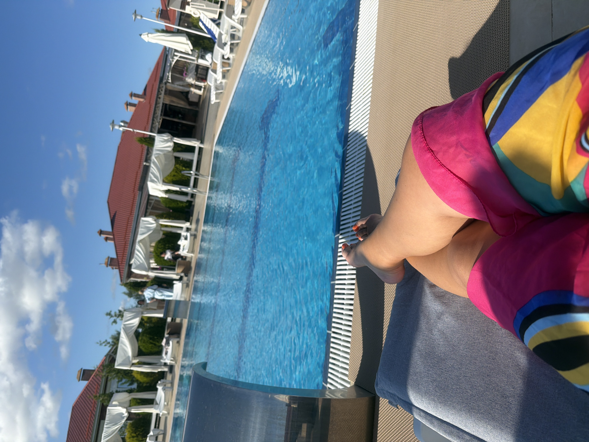 The image depicts a close-up view of a person's legs and torso, resting on a lounge chair beside a swimming pool. The person is wearing colorful shorts with a bright pink, yellow, blue, and black geometric pattern. Their feet, with orange-painted toenails, are visible at the bottom of the frame. The lounge chair is covered with a gray cushion.
Beyond the lounge chair is a bright blue swimming pool with lane markings visible beneath the surface. The pool is bordered by a white grate system. In the background, several white cabanas with draped fabric are set up, and beyond that, buildings with red roofs are visible. There are also some green bushes and trees. A few people are seen in the background, one in a light blue robe, and others standing near the cabanas and buildings. The sky is blue with scattered white clouds.
The scene appears to be an outdoor leisure area, likely a resort or hotel, given the presence of the pool, lounge chairs, and cabanas. The location is identified as Laskino, Russia. The lighting suggests it is daytime, with sunlight casting shadows. The perspective is from the viewpoint of someone lying on the lounge chair, looking towards the pool and the surrounding area.