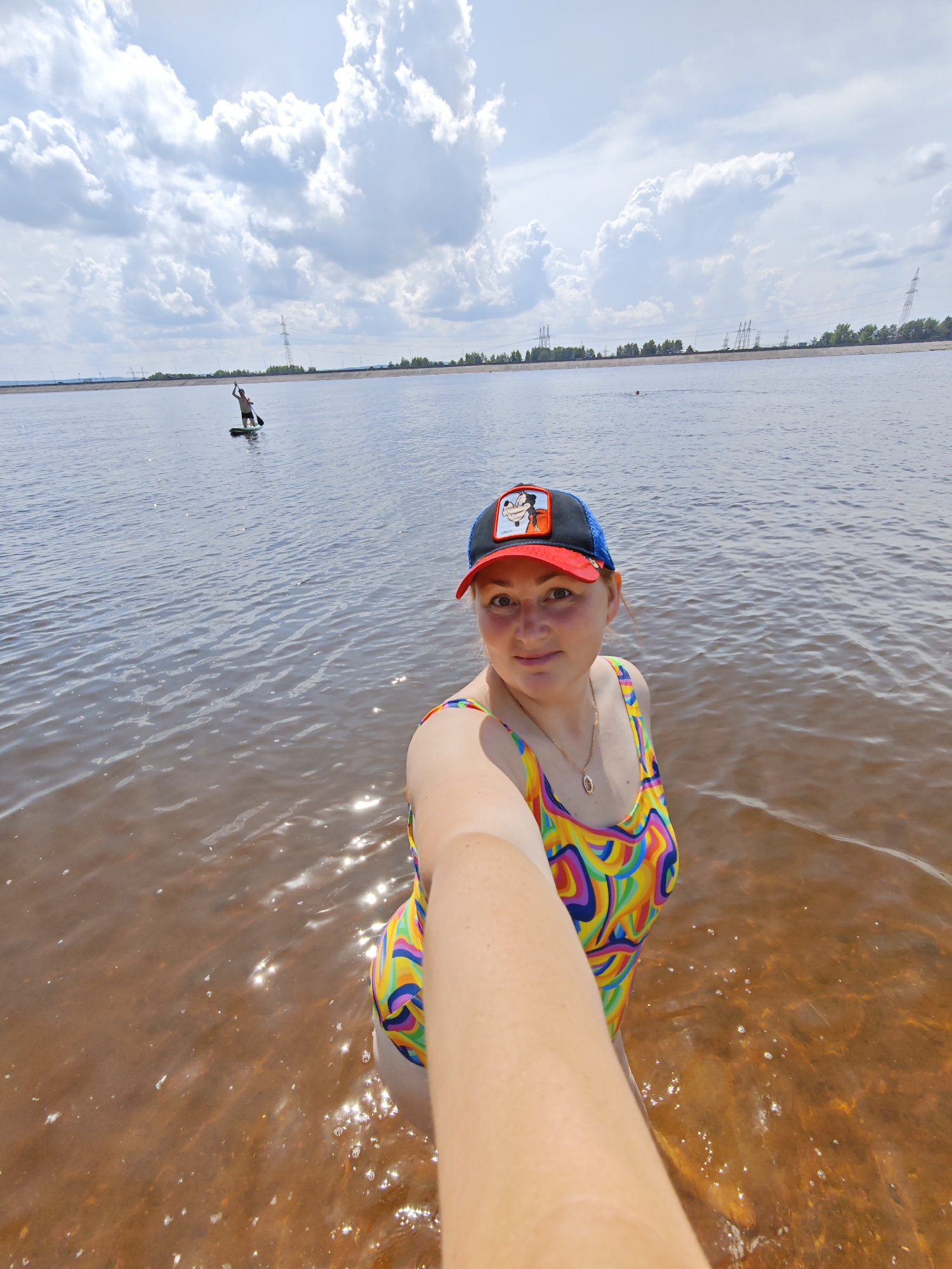 In a wide shot, a woman stands in shallow, reddish-brown water, holding a camera to take a selfie. She is wearing a colorful, swirling-patterned swimsuit and a blue and red baseball cap with a cartoon character on it. The sun is shining brightly, creating a shimmering effect on the water's surface and casting shadows. The sky is a bright blue with large, fluffy white clouds. In the background, a person is paddleboarding on the vast body of water. Further in the distance, a tree line marks the shore, with several tall power line towers visible behind it. The scene appears to be a sunny day, likely in the afternoon, at a lake or river near Noviy, Russia.