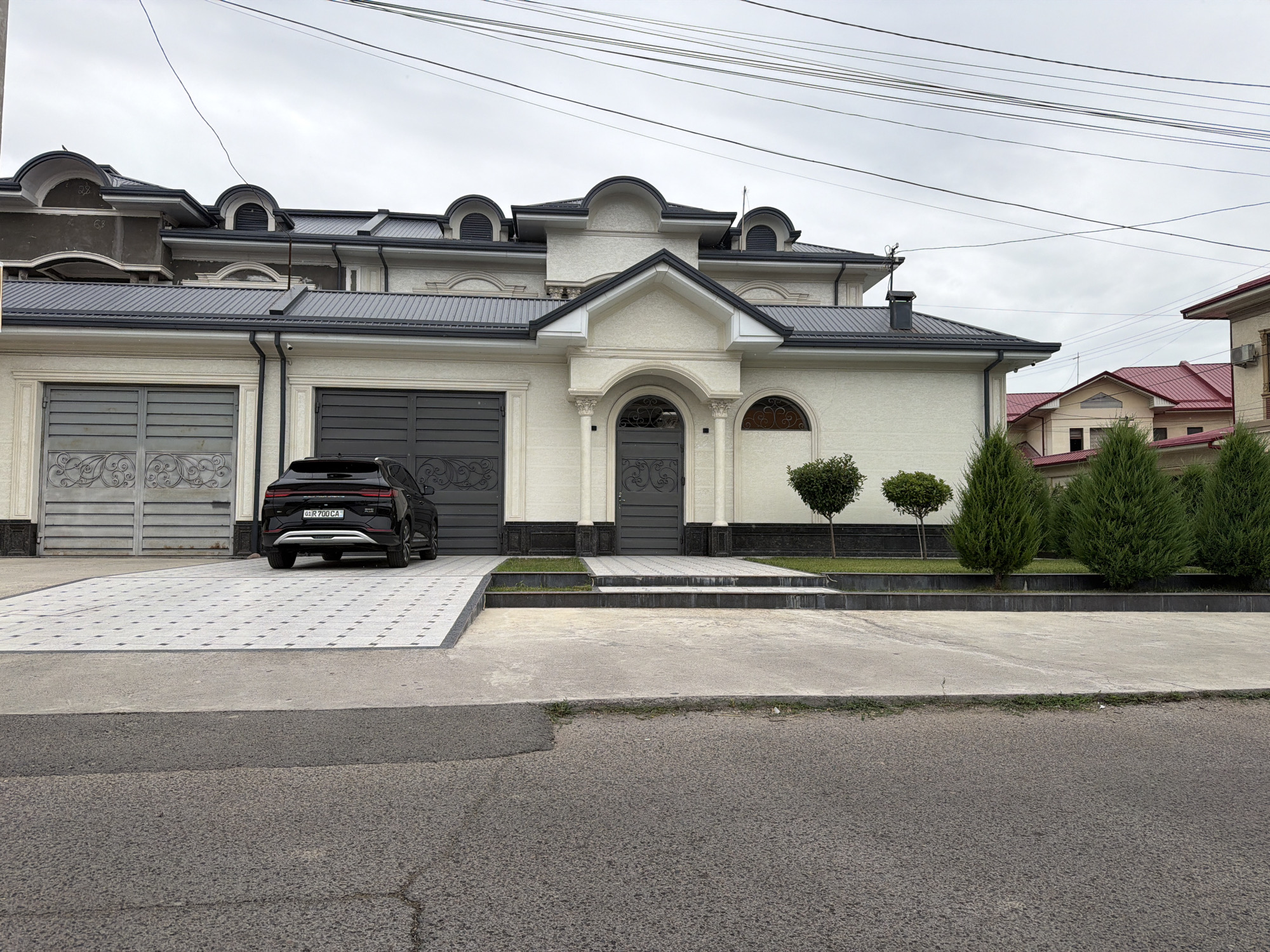 The image shows a large, modern house with a dark gray metal roof, cream-colored stucco exterior, and decorative architectural elements. The house features multiple dormer windows with arched tops and dark louvers. There are two large garage doors on the left side of the house, one with a gray, ornate metal design and the other a plain dark gray. A black SUV with a license plate reading "DJR 700 CA" is parked in front of the driveway. The driveway is paved with light-colored tiles arranged in a grid pattern. In front of the house, there is a low, dark gray border with a small strip of grass, followed by a raised concrete sidewalk. To the right of the entrance, there are three small, manicured trees and several taller evergreen trees. In the background, other houses with red tile roofs are visible. Overhead, multiple power lines crisscross the cloudy sky. The scene appears to be in a residential area in Tashkent, Uzbekistan, indicated by the style of architecture and the car's license plate.