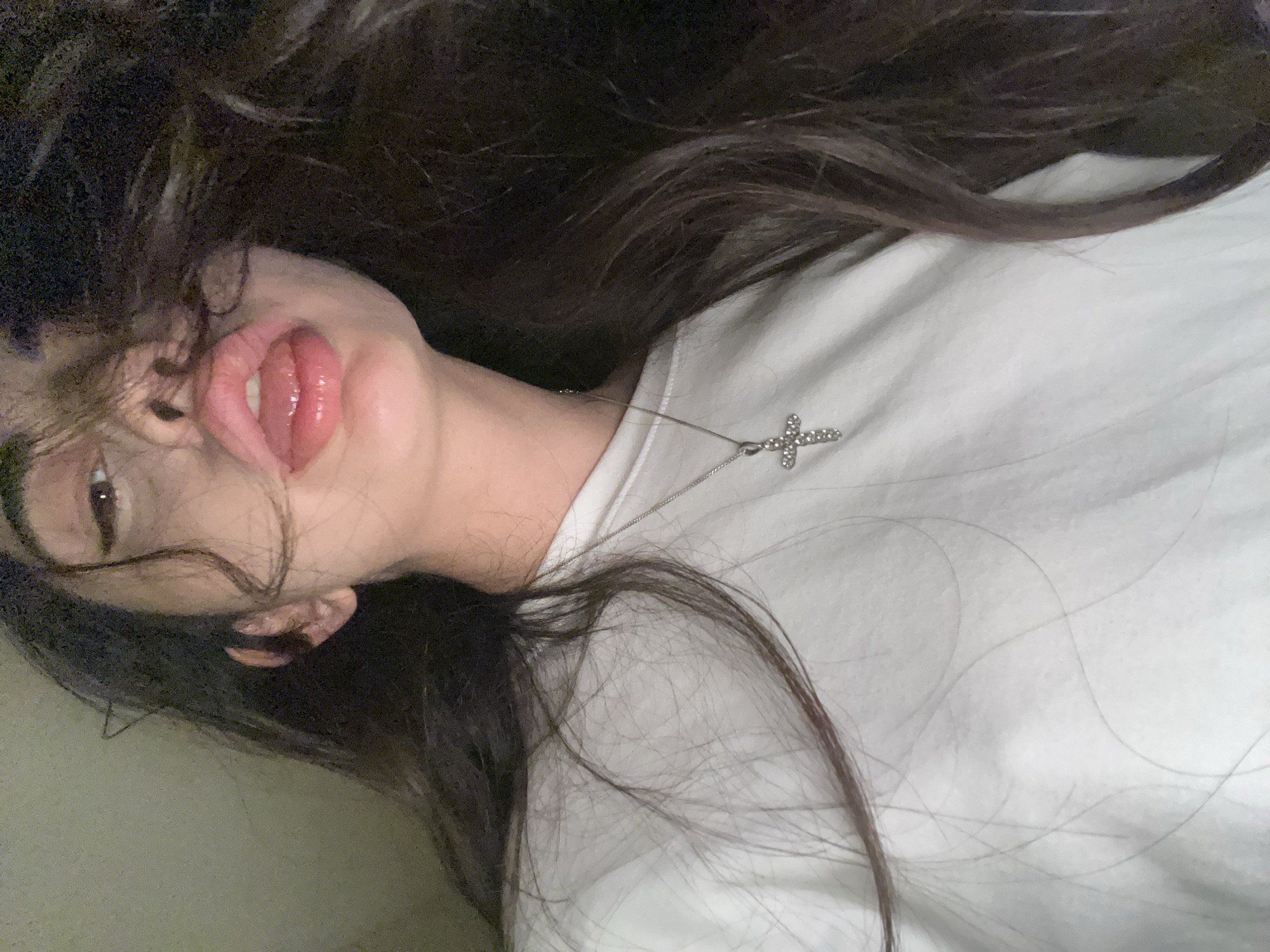 The image shows a close-up of a young woman's face and upper chest. She has long, dark hair that is partially obscuring her face. She is wearing a white sweatshirt with a silver cross necklace. Her lips are slightly parted and appear to be glossy. The background is dark and out of focus. The image was taken indoors, likely in a residential setting.