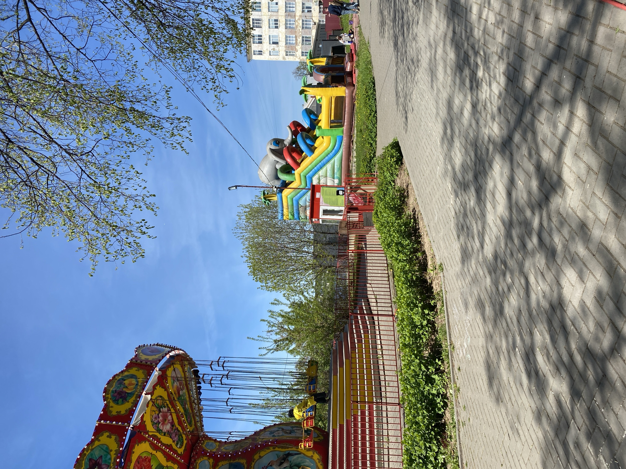 The image depicts an outdoor amusement park scene in Izhevsk, Russia, featuring several attractions. In the foreground, the top portion of a carousel swing ride is visible, with its ornate, colorful decorations. Moving towards the middle distance, a large, inflatable bouncy castle stands adjacent to a paved walkway. The walkway is lined with a low fence and some greenery. In the background, an apartment building can be seen. The sky is blue, with a few wispy clouds. There is a person on the carousel swing ride. There are no people near the bouncy castle.