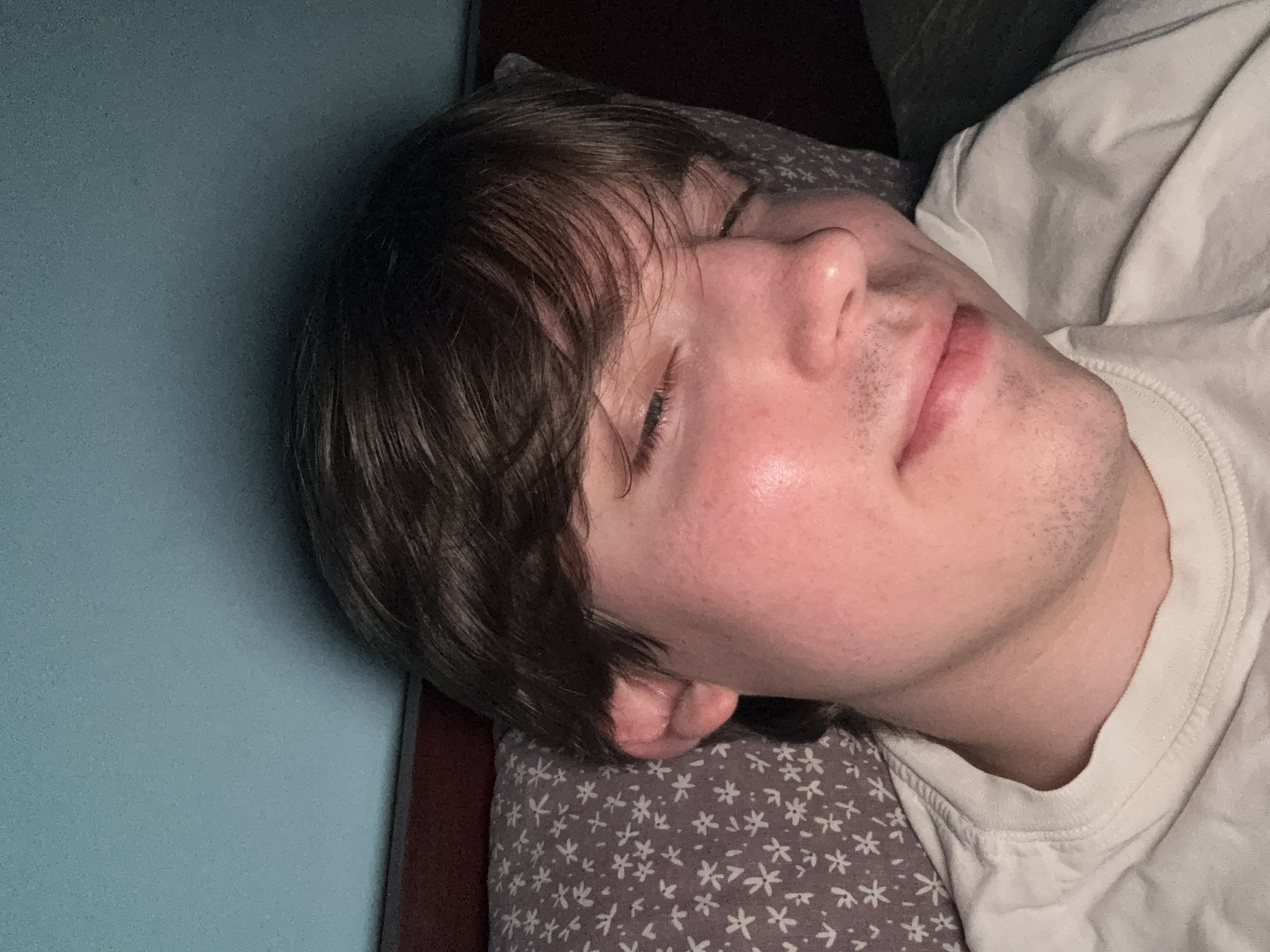 The image shows a close-up, head-and-shoulders shot of a young man with brown hair and closed eyes, resting his head on a patterned pillow. He is wearing a white t-shirt. The background consists of a red headboard and a turquoise wall. There is no indication of the location being in the city of Dnipro, Ukraine.