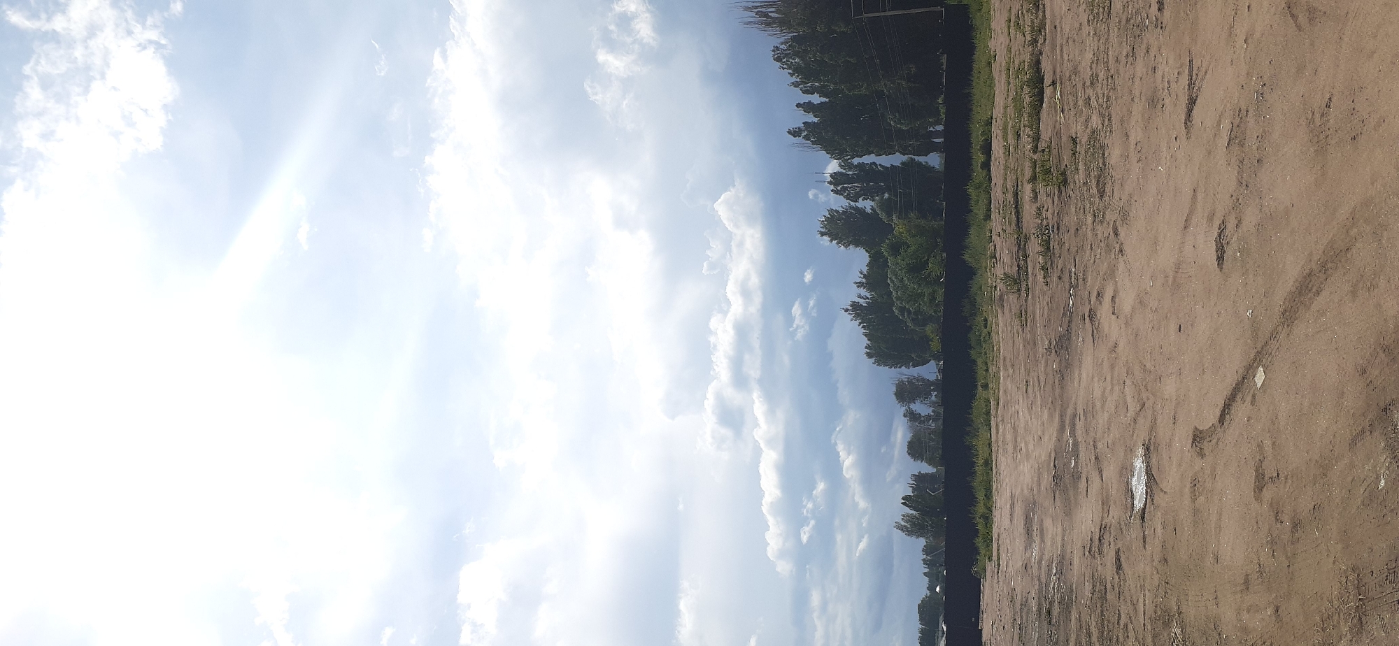 The image depicts an outdoor scene on a partly cloudy day, likely in Chok Tal, Kyrgyzstan. The foreground consists of a dirt ground with tire tracks, suggesting it may be a pathway or an undeveloped area. In the midground, a black fence runs horizontally across the image, separating the foreground from a strip of green grass and vegetation. Behind the fence, a line of trees, varying in height and density, forms a natural barrier. The sky occupies the upper portion of the image, with large, white clouds and streaks of sunlight indicating a bright, though not entirely clear, day. There are no people or animals visible in the frame.