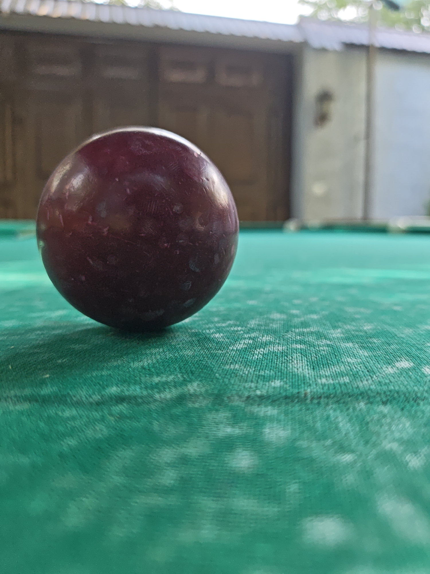 A close-up, low-angle shot shows a dark red billiard ball resting on a green felt surface. The ball is slightly to the left of the frame's center, with the felt extending diagonally towards the lower right. The background is out of focus, showing a blurred structure that appears to be an outdoor building with a corrugated roof and a large wooden door. The scene is set outdoors, likely in Yakkatut, Uzbekistan, given the architectural style. No people are visible.