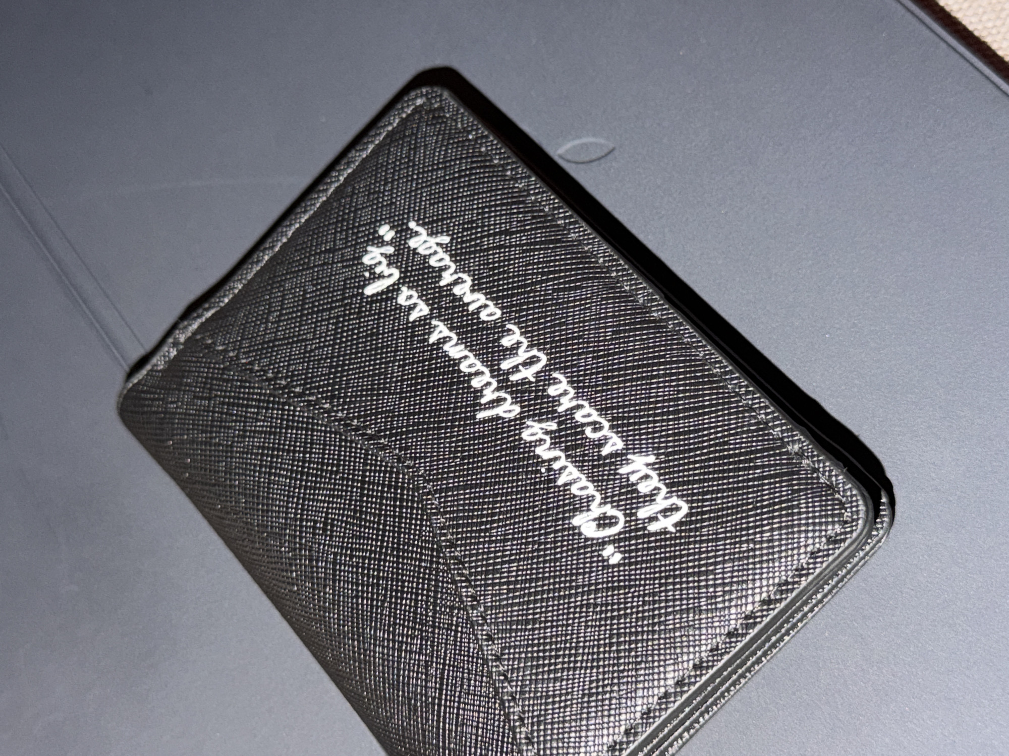 The image displays a dark-colored wallet or cardholder resting on a flat, dark grey surface. The object is rectangular and features a textured, cross-hatch pattern, consistent with Saffiano leather.
Prominently featured on the front of the wallet is white cursive text. The inscription reads: "Chasing dreams is to live The average." The words "Chasing dreams is to live" are on the first line, followed by "The average" on the second line.
The setting appears to be indoors, under artificial lighting, with the wallet positioned diagonally across the frame. There are no people, discernible activities, or specific location cues. The background surface is plain and unadorned.