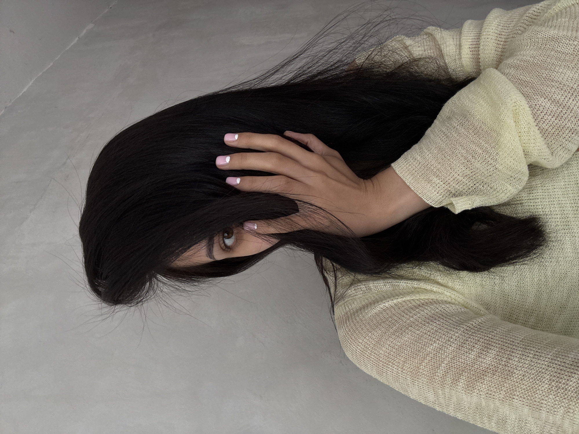 The image presents a close-up, diagonal view of a person with long, dark hair, set against a plain, light grey background that resembles a wall or floor. The individual's face is largely obscured by their voluminous, dark hair, with one brown eye peering out from behind it, creating a sense of partial concealment.
A hand is gently placed amidst the hair, showcasing neatly manicured nails painted in a soft pink hue, each adorned with a distinct white half-moon design near the cuticle. The person is wearing a light yellow or cream-colored, loosely-knit garment, possibly a sweater or top, visible on the right side of the frame. The textile's open, mesh-like weave is clearly distinguishable.
The composition highlights the textures of the hair, skin, and fabric against the neutral background. The visible eye provides a focal point, suggesting a direct yet partially hidden gaze. The lighting appears soft and diffused, indicating an indoor setting. There are no identifiable objects or text in the background to provide further context about the location or time of day. The overall impression is one of introspection, mystery, or perhaps a playful peek from behind a curtain of hair.