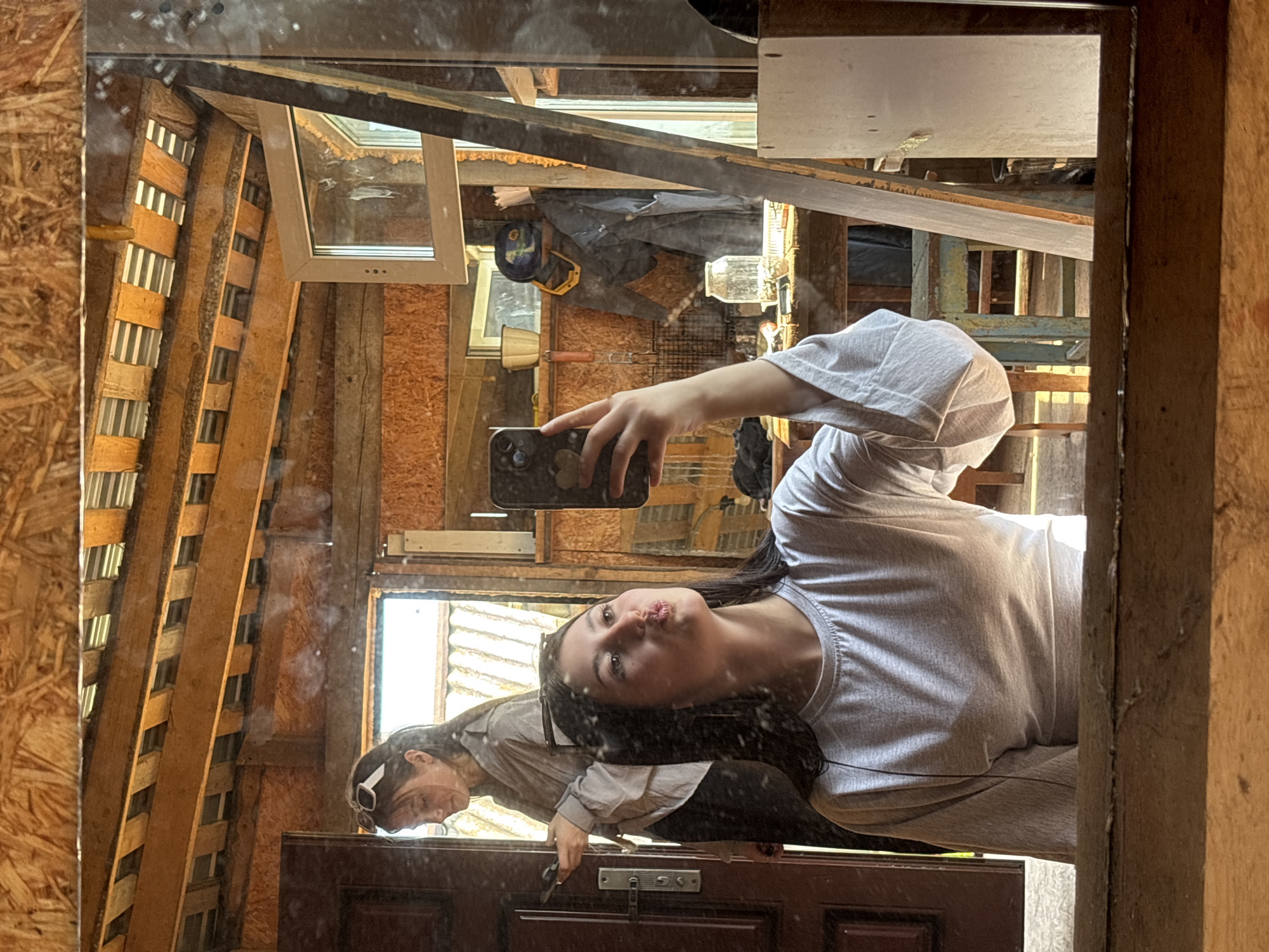 The image depicts a reflection in a mirror, showing a young woman taking a selfie. She is wearing a grey t-shirt and holding a smartphone. In the mirror's reflection, another person is visible. The setting appears to be a cluttered workspace or shed. Various tools, lumber, and construction materials are visible. The location, Фоки, Russia, is not determinable from the visual elements in the image.