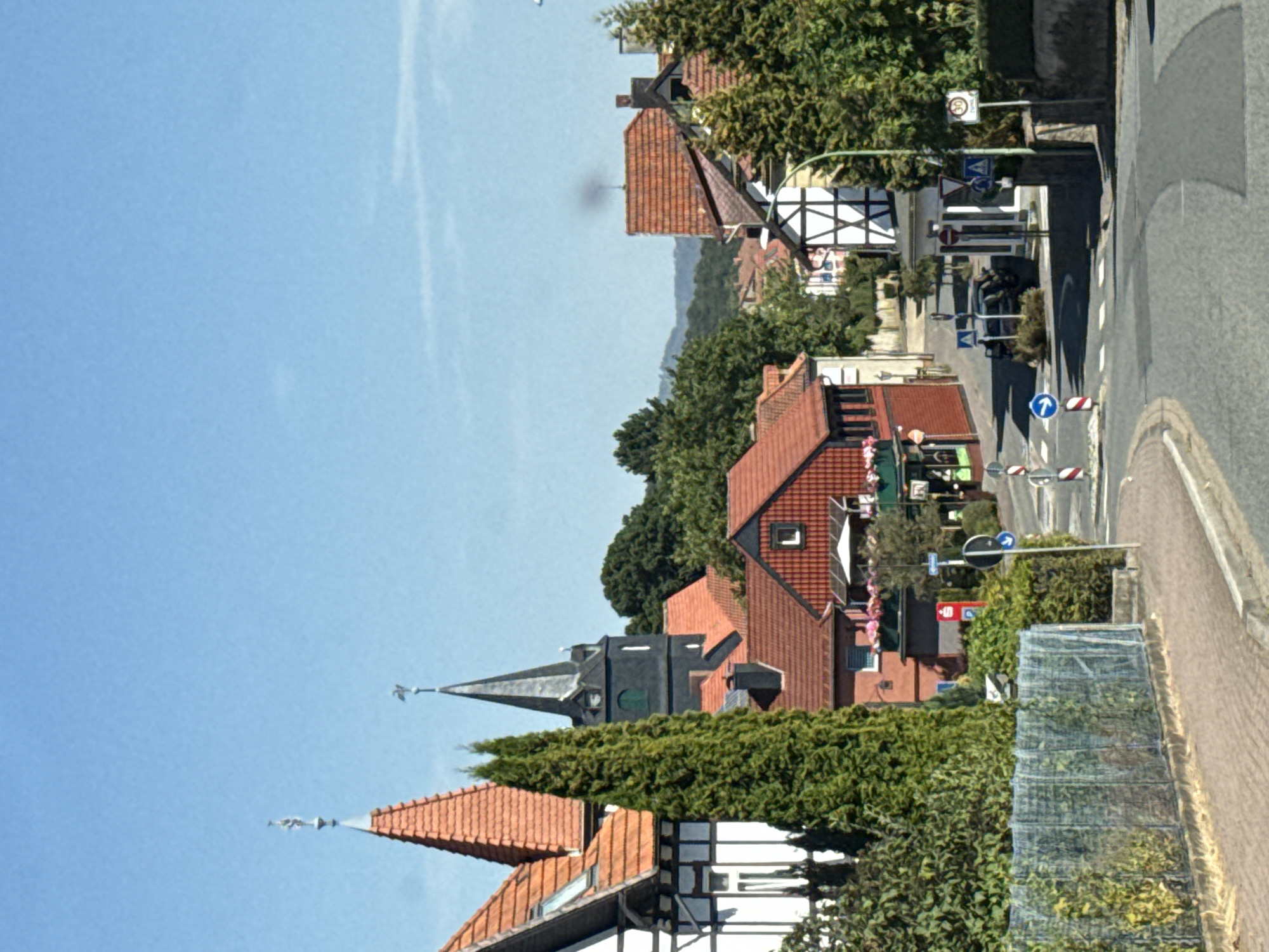 This is a street-level photograph of the town of Bad Sachsa, Germany. The scene features traditional German architecture with half-timbered houses and red-tiled roofs, along with a church spire. The sky is clear and blue, indicating a sunny day. The road in the foreground is paved and curves to the right, with a sidewalk and a metal fence lining the left side. Various road signs are visible, including a speed limit sign and directional signs. Green trees and bushes are interspersed throughout the scene, adding to the natural beauty of the town. The overall impression is one of a peaceful and picturesque German village.