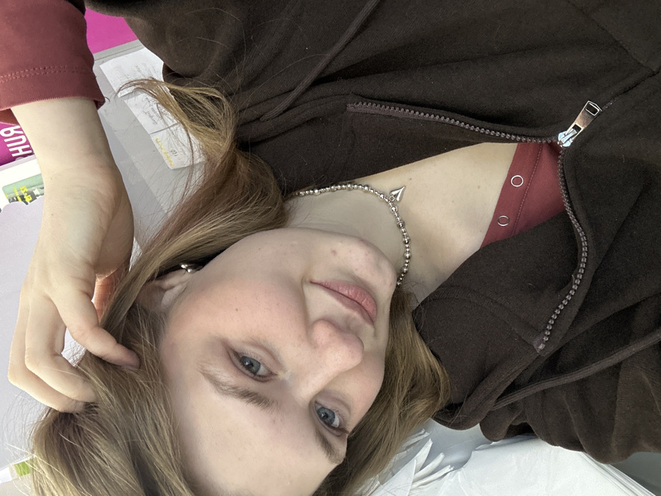 The image displays a close-up portrait of a young, light-skinned female in an indoor setting within Chaikovsky, Russia. The subject, with blonde hair and blue eyes, is captured with a slight smile, looking directly towards the viewer. Her left hand is positioned with fingers lightly touching her hair above her right ear. She is wearing a dark brown zippered hoodie, partially unzipped, revealing a reddish-brown garment beneath which exhibits two small circular fastenings. Accessories include small silver-colored hoop earrings and a silver-colored beaded necklace featuring a prominent, pointed heart-shaped pendant. The background, blurred, features a white object or stack of items on the left. To the upper right, a portion of a sign with Cyrillic text, specifically the letters "-ИЯ" on a pink background, is visible, alongside a light-colored surface with faint yellow writing and the numeral "110". The environment suggests a casual, possibly retail or administrative indoor space.