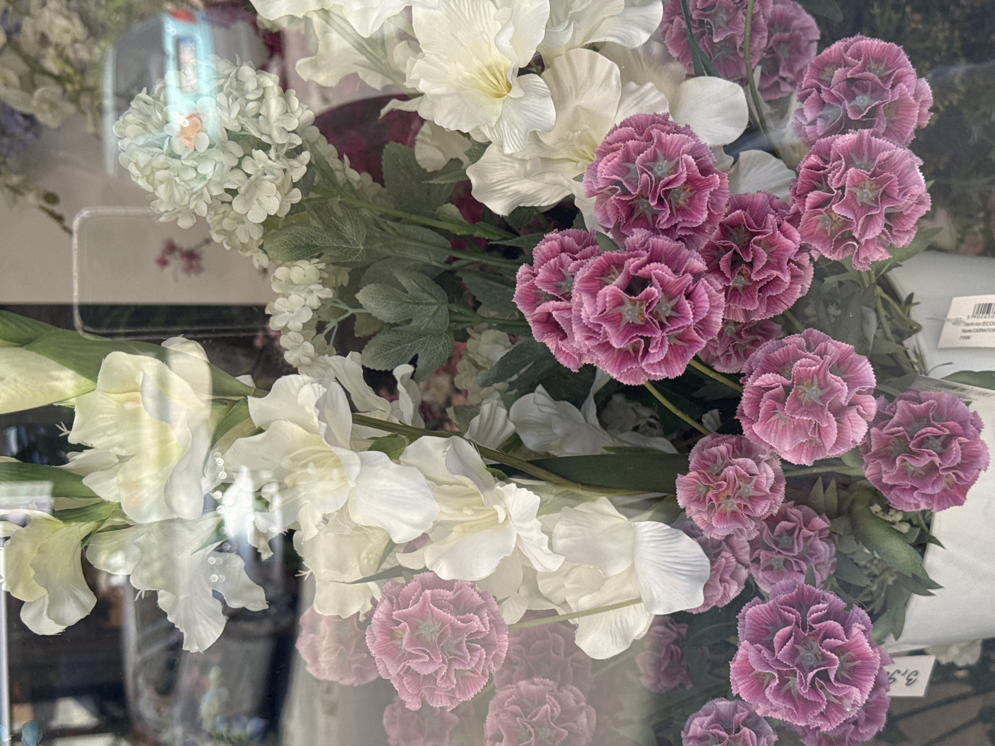 The image shows a close-up view of a floral arrangement, likely displayed in a shop window, with a blurry reflection of a street scene and vehicles in the background. The flowers consist of white gladiolus-like blooms and clusters of pink carnations, all appearing to be artificial. The arrangement is in a white pot or container. A small price tag is visible on the pot. The reflection shows a street with cars, indicating an outdoor urban environment. There are no people clearly visible. The overall impression is of a commercial display. The location is identified as Budva, Montenegro.