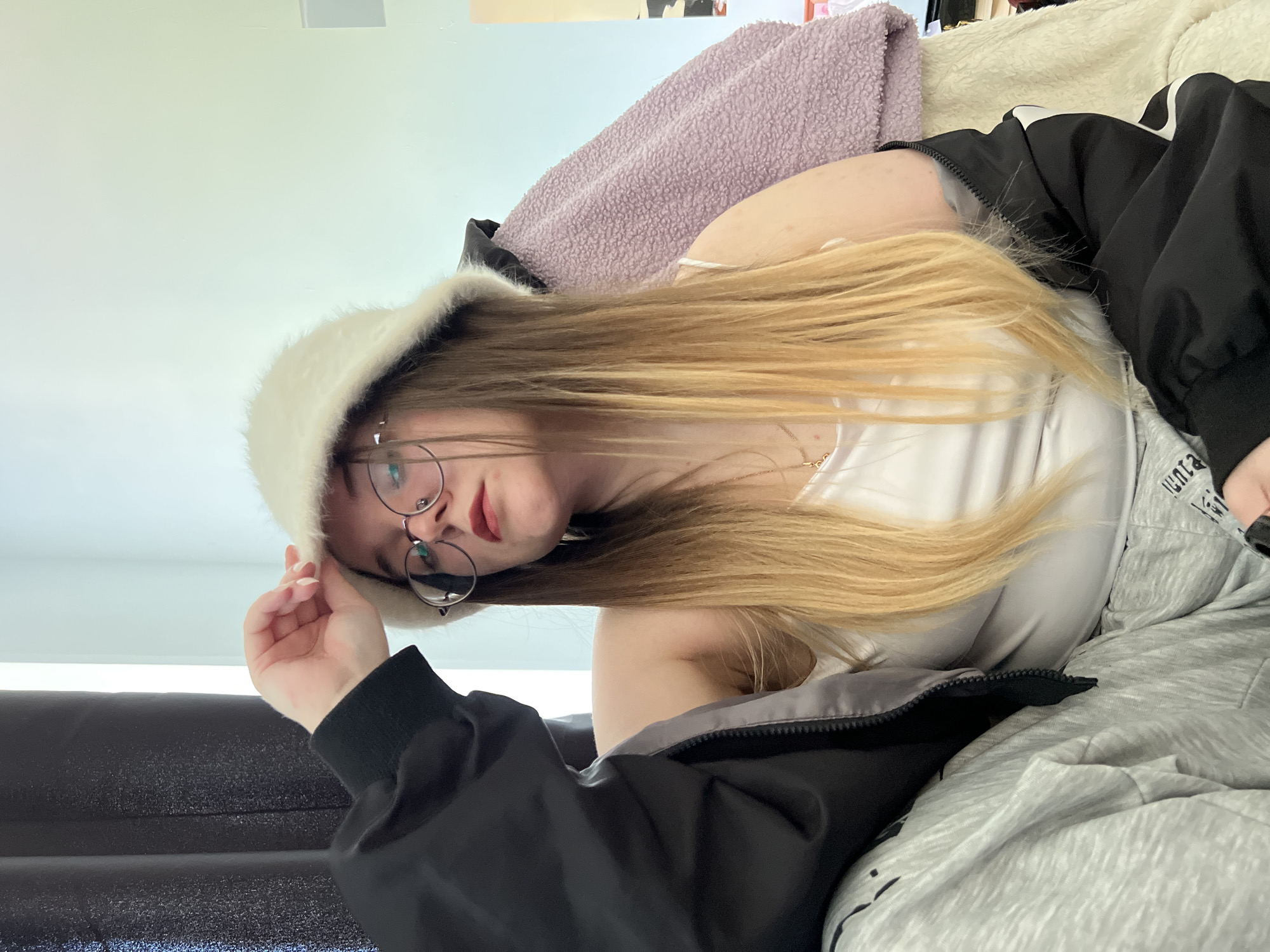 A young woman with long blonde hair, wearing round glasses and a white furry hat, is reclining on a gray and white patterned surface. She is also wearing a white tank top and a black jacket. A lavender blanket is behind her. The photo appears to be taken indoors, and the scene's location is likely not Pushchino, Russia.