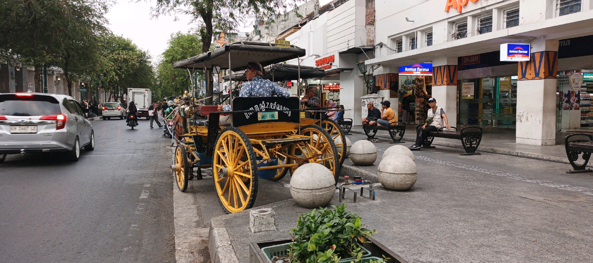This image captures a street scene in Yogyakarta, Indonesia, likely in the tourist area of Malioboro Street, characterized by its horse-drawn carriages (Andong). In the foreground, a yellow and black Andong is parked on the side of the road, its driver, a man wearing a blue patterned shirt, visible at the front. Several large, spherical concrete bollards are arranged along the sidewalk, with a man in a vest and cap sitting on a dark bench nearby. Behind the Andong, traffic moves along the street, including a silver car, a white truck, and several motorcycles. People are seen walking and riding on the motorcycles. Buildings with storefronts, signage for "KIMIA FARMA," and awnings line the opposite side of the street. The sky is overcast, and the street appears wet, suggesting recent rain. The overall atmosphere is that of a bustling urban environment.