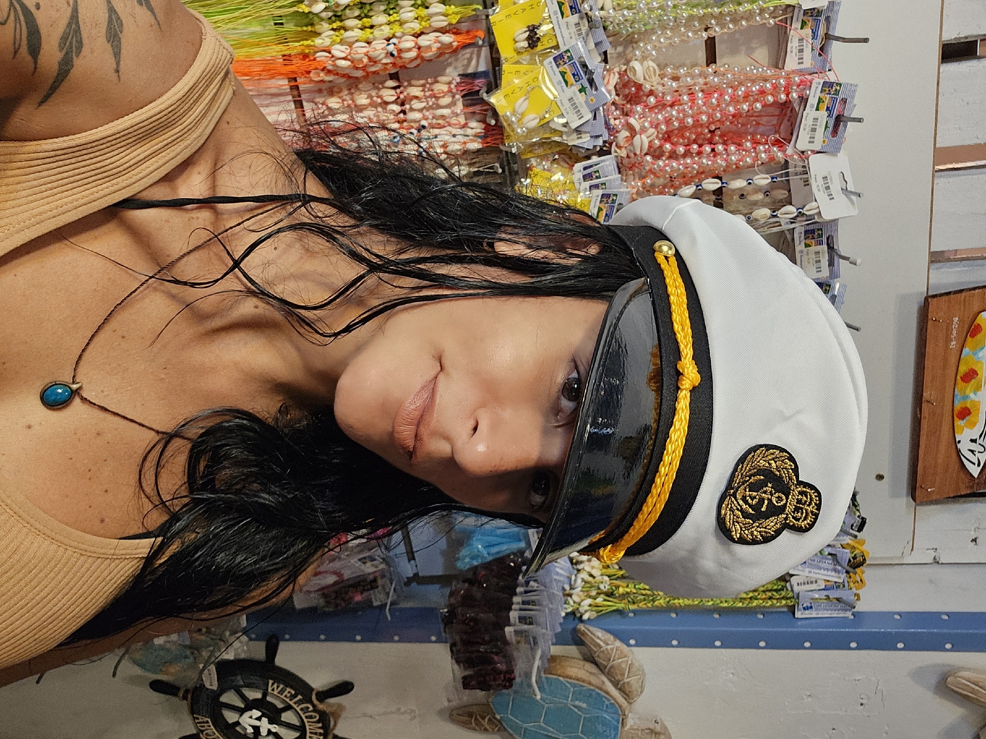 A person with dark hair, wearing a beige top and a captain's hat, is posing for a photo in a shop. The background features shelves displaying various items, including colorful necklaces and beaded accessories. In the lower part of the frame, there are nautical-themed decorations, such as a ship's wheel and a turtle figure. Based on the items in the background and the general aesthetic, the location could be a souvenir shop in Armação Dos Búzios, Brazil, given that it is a coastal town known for its beaches and tourism. The person is looking directly at the camera.