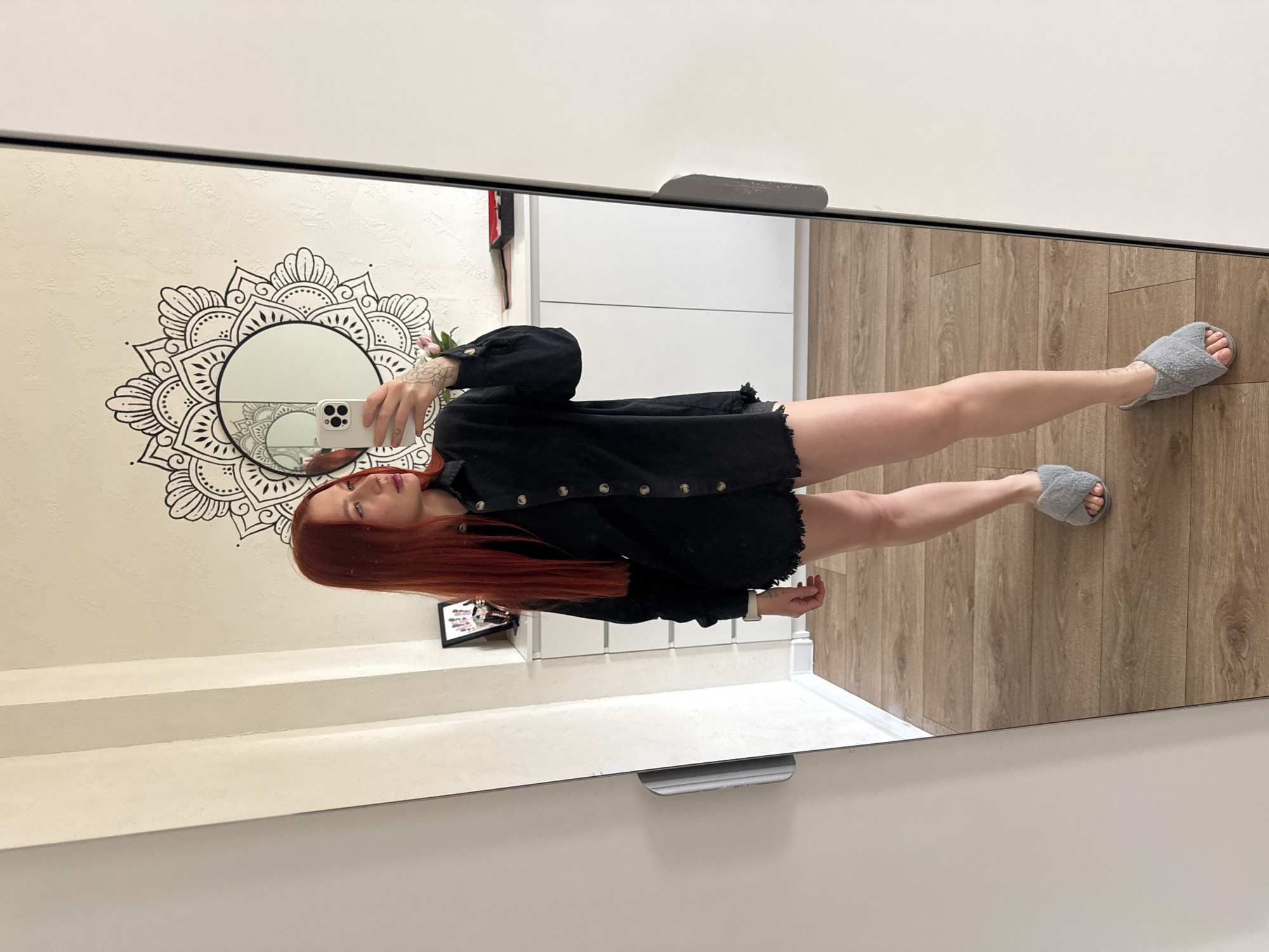 The image is a reflection in a large mirror, likely in an interior space. A woman with long, red hair is holding a smartphone, presumably taking the photo. She is wearing a black, button-down shirt that appears slightly oversized and frayed at the bottom. The lower half of her body is visible, revealing her legs and feet clad in gray, fuzzy slippers.
The backdrop includes a wall decorated with a large mandala design around a smaller, circular mirror. To the right of the woman, in the reflection, is a light wood floor. In the background, there's a white cabinet or shelving unit.
The scene suggests a personal space, possibly a bedroom or dressing area, given the presence of the mirror, decorative wall art, and relaxed attire. The location in Rostov-on-Don, Russia, is not evident from the visual details within the image.