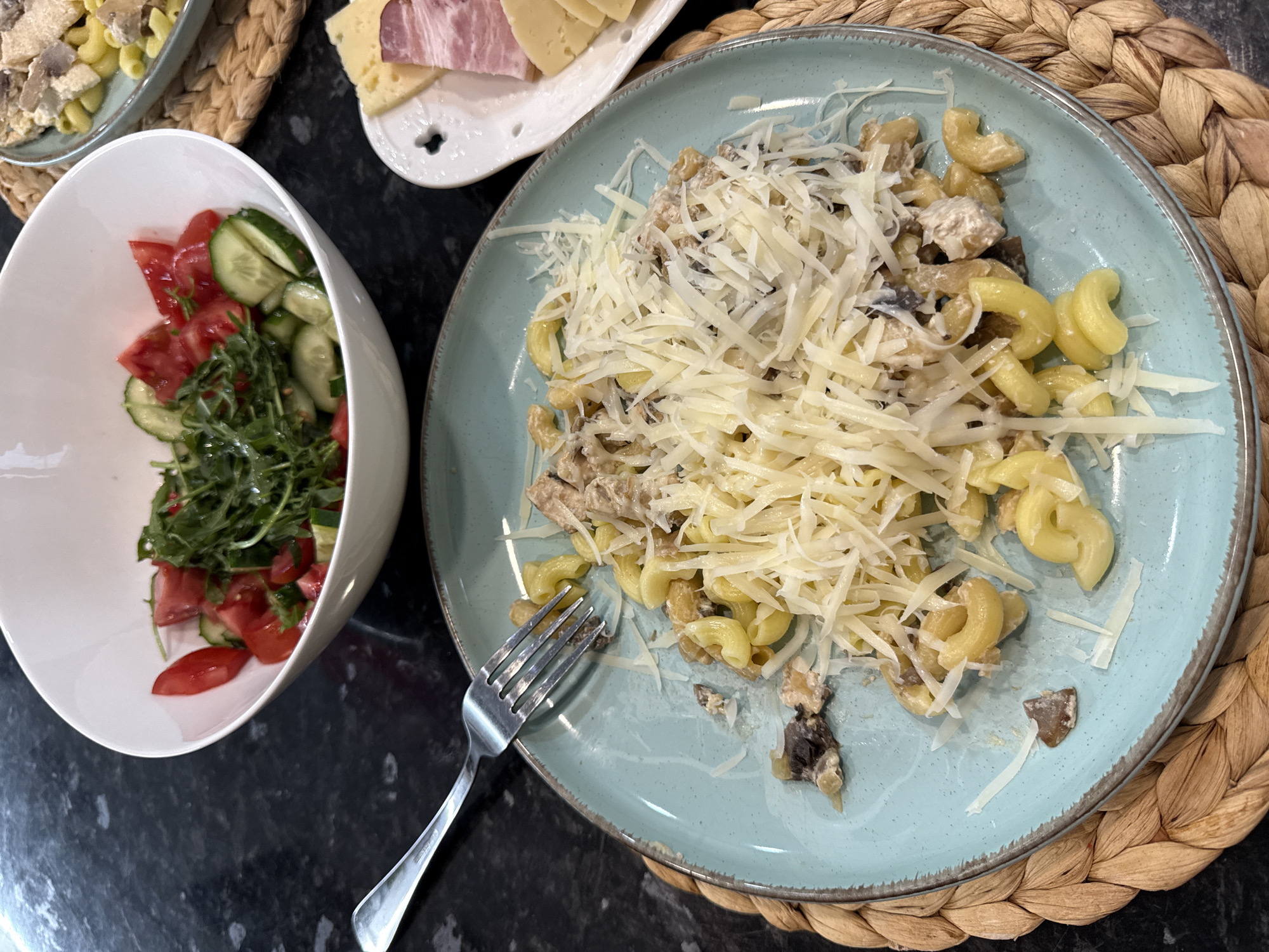 A meal is presented on a dark, speckled countertop. In the foreground, a light blue plate holds pasta topped with shredded cheese and what appears to be chicken and mushrooms in a creamy sauce. A fork rests on the edge of the plate. To the left, a white bowl contains a salad of chopped tomatoes, cucumbers, and greens. To the right, a small white plate displays slices of cheese and ham. The scene is lit from above, casting soft shadows. No specific location in Sochi, Russia, is discernible.