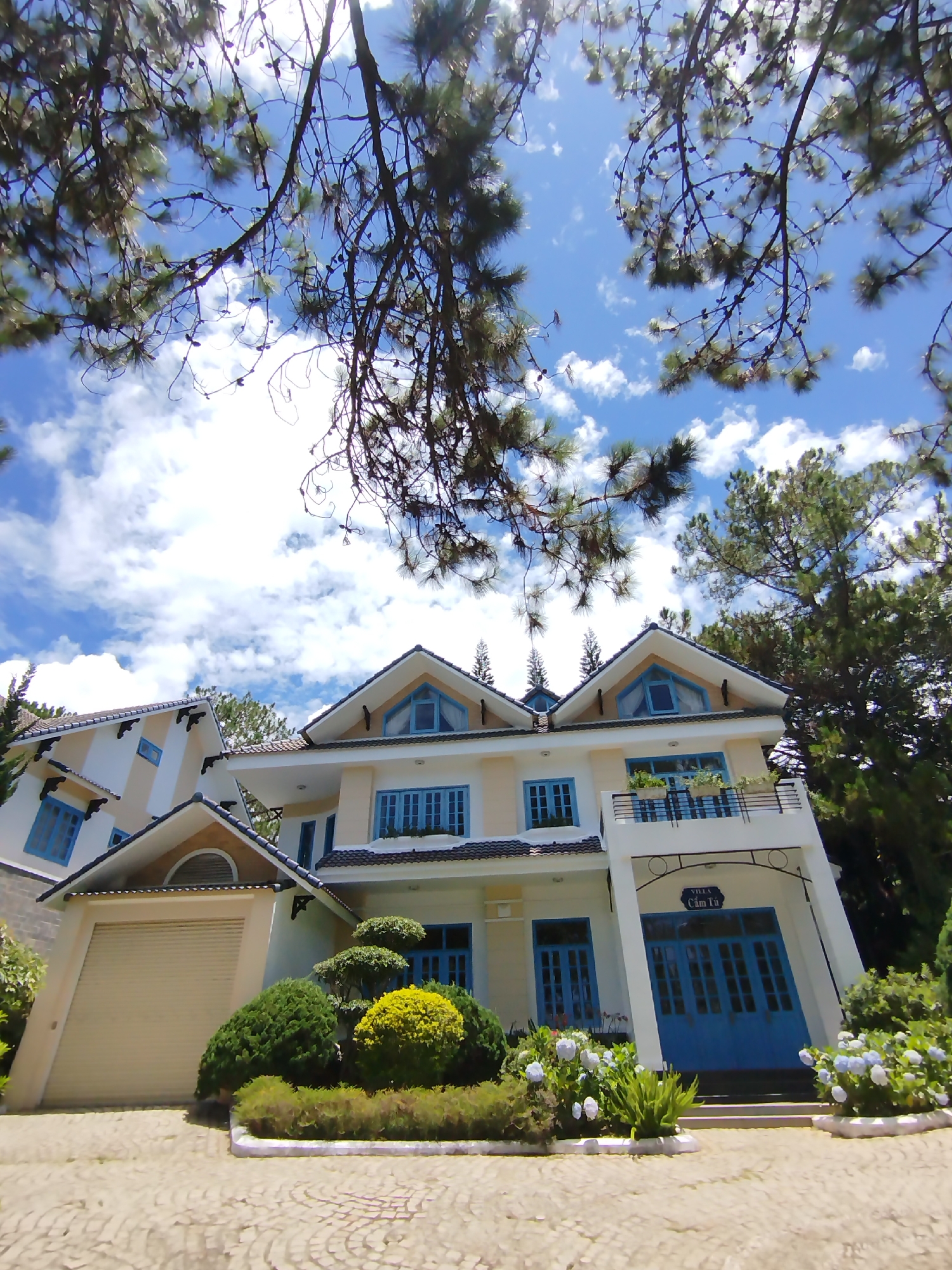 The image captures a two-story house with a light yellow exterior and blue accents around the windows and doors. A detached garage is connected to the main house via a covered entryway. The house is surrounded by lush greenery, including various shrubs and hydrangeas. Above, tree branches frame a view of a blue sky dotted with clouds. A sign near the entrance reads "Villa Cẩm Tú," indicating the location in the city of Lâm Viên - Đà Lạt, Vietnam. The pavement in front of the house appears to be patterned brick.