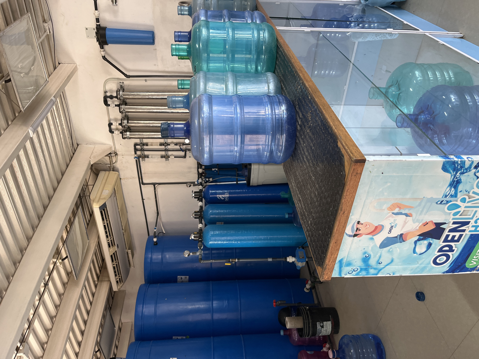 The image shows an indoor water purification and bottling facility in Juan Griego, Venezuela. Large blue plastic water jugs are stacked for filling, while others are displayed on a glass-topped counter with a promotional advertisement. A series of blue cylindrical water filters and stainless-steel filtration units are mounted on the wall, connected by black pipes, illustrating the purification process. An air conditioning unit is visible on the ceiling. A water pump and additional water jugs are on the floor.