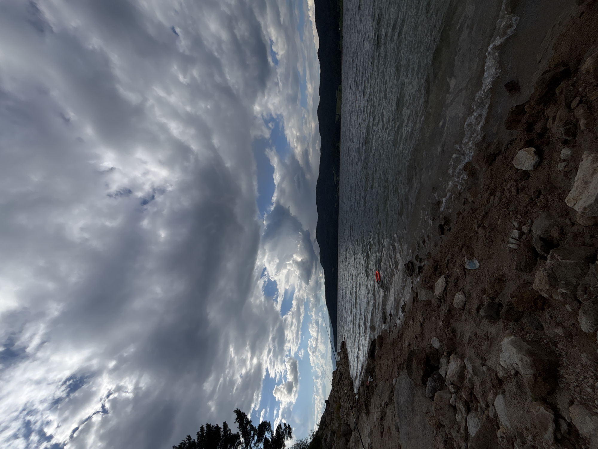 A wide-angle, low-angle shot captures a rocky shoreline bordering a large body of water under a dramatic, cloudy sky. The foreground is dominated by a rough, uneven shore littered with rocks and dirt, with a few pieces of debris visible. A dark, coniferous tree stands on the left side of the frame, partially obscuring the view of the sky and mountains in the distance.
The water is dark and choppy, with small waves lapping at the shore. In the middle ground, a bright orange object, possibly a buoy or a life preserver, floats on the water's surface. Across the water, a dark, forested mountain range stretches across the horizon, its silhouette softened by the distance and atmospheric conditions.
The sky is filled with a dynamic display of clouds. Large, dark, and heavy clouds dominate the upper half of the frame, casting a moody atmosphere. However, breaks in the clouds reveal patches of bright blue sky and shafts of sunlight, creating a sense of contrast and impending weather. The overall impression is one of natural beauty with a hint of wildness, perhaps taken during a moment of transition between fair and stormy weather. The location is an unspecified natural setting.