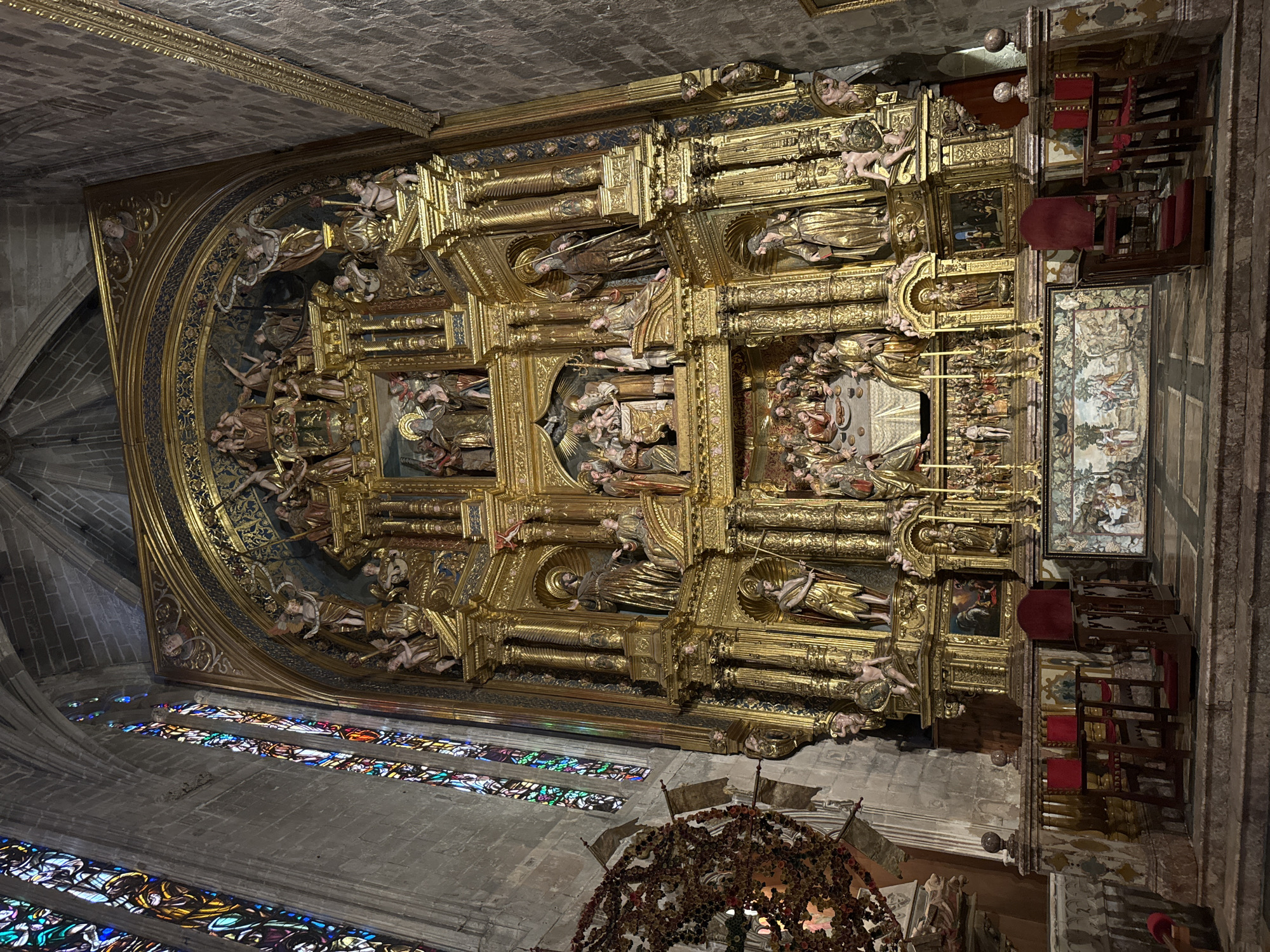 The image depicts the gilded altarpiece of the La Seu Cathedral in Palma de Mallorca, Spain. The altarpiece is a large, ornate structure adorned with numerous sculpted figures and decorative elements, primarily in gold. Stained glass windows are visible on the left side of the image, casting colorful light into the cathedral. In the foreground, there are several wooden pews with red cushions. The scene is static and appears to be an interior shot of a religious building.