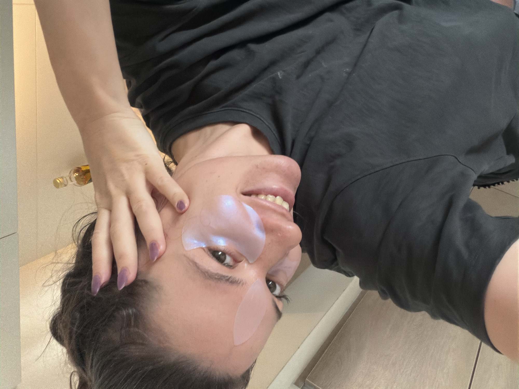 A person, likely female, is posing for a selfie indoors. They are wearing under-eye patches and a dark t-shirt, with their hair pulled back. One hand is near their face, with visible fingernails painted a purple-ish hue. A bottle is visible in the background. Given the tile pattern in the background, it is possible this image was taken in a kitchen or bathroom. There is no information to suggest that this photo was taken in Belgrade, Serbia.