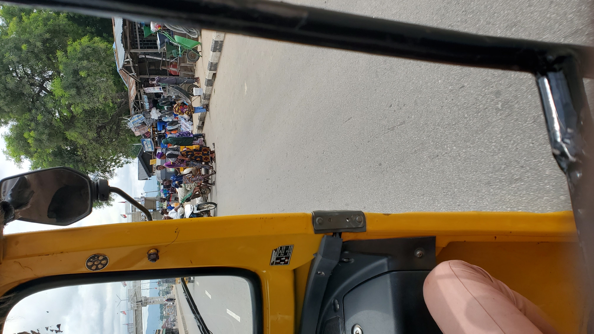 The image depicts a street scene in Jalingo, Nigeria, viewed from the interior of a yellow auto-rickshaw. In the foreground, the yellow body of the auto-rickshaw is visible, including a side mirror and part of the dashboard. To the left, a person's legs, clad in light pink trousers, are visible. The street is paved and lined with shops and stalls on the right side of the image. A crowd of people, including men and women in traditional attire, are gathered on the sidewalk and street, some interacting with vendors. Several motorcycles, carts, and a bicycle are also present, indicating a busy market or thoroughfare. The background features trees, buildings, and a bridge with flags, suggesting a populated urban environment. The sky is overcast, and the overall atmosphere appears to be that of a typical day in a Nigerian city.