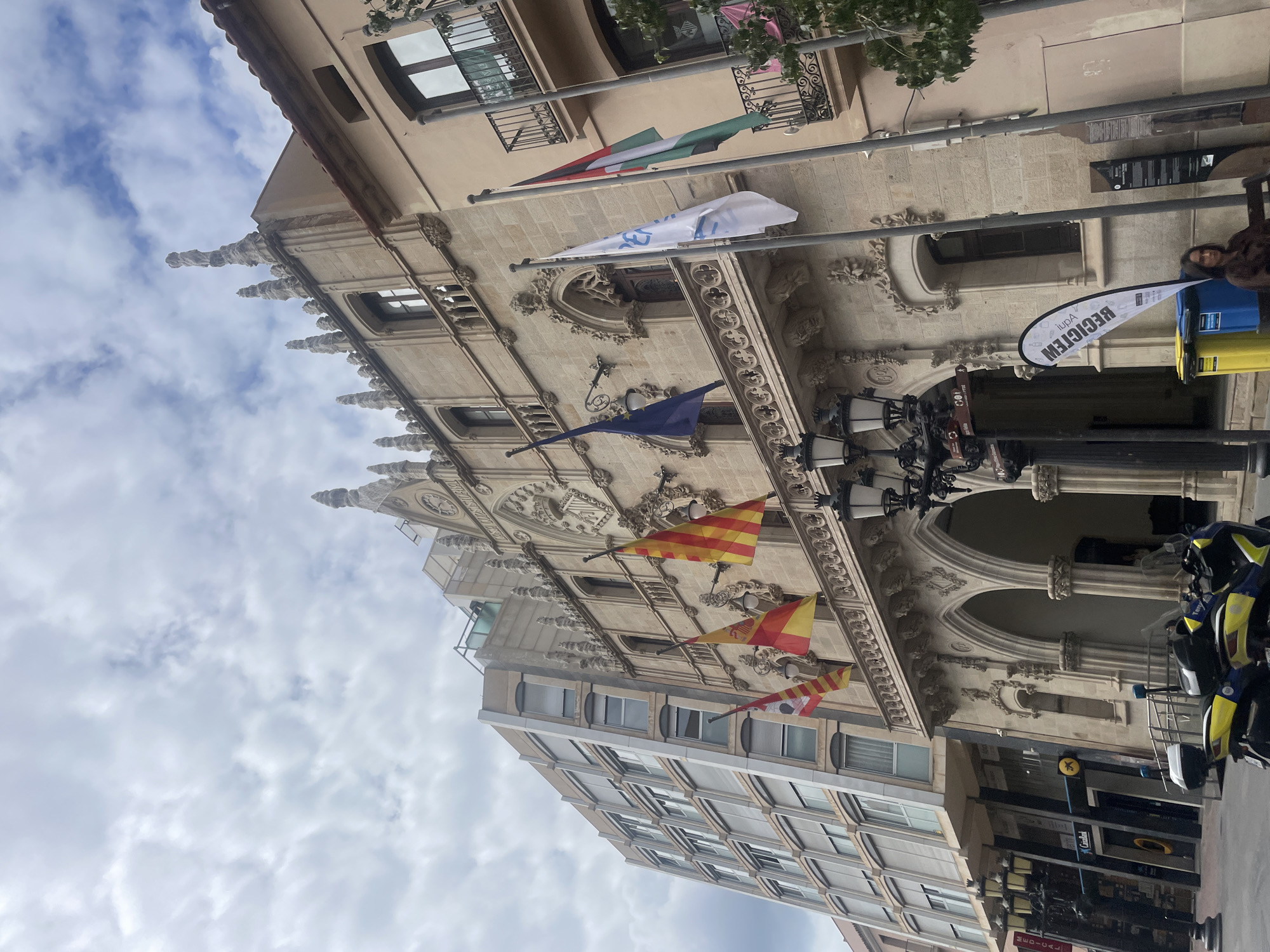 The image shows a low-angle view of a building in Terrassa, Spain. The architecture features both ornate, historical elements (like pointed arches, decorative stonework, and multiple flags, including what appear to be Catalan flags) and a more modern, box-shaped addition. Street elements are visible, including streetlights, a recycling bin, and a motorcycle. The sky is cloudy. A person is partially visible on the right.