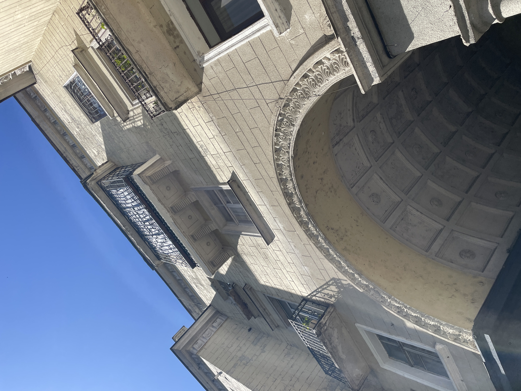 The image captures the architecture of a building in Tbilisi, Georgia, from a low-angle perspective pointing upward. The building is constructed of stone or concrete blocks and exhibits various architectural details, including balconies with wrought iron railings, windows with ornamental frames, and a large arched opening decorated with relief patterns along its curve. The archway reveals a coffered ceiling. The building's facade is weathered and shows signs of age. A clear, bright blue sky is visible on the left side of the frame. The lighting indicates a sunny day.
