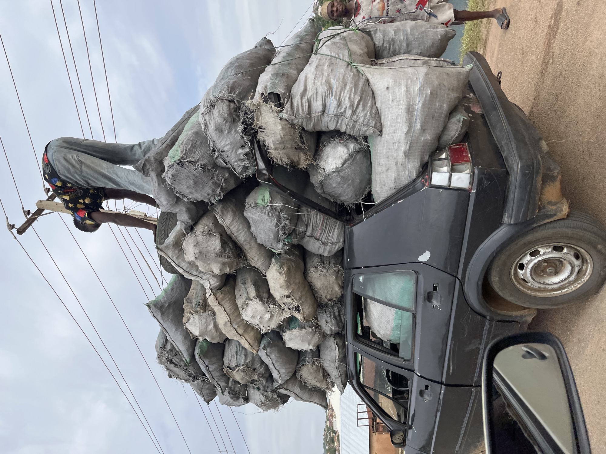 The image depicts a heavily overloaded black hatchback car on a street in Jos, Nigeria. The vehicle is stacked precariously high with numerous gray sacks, likely containing charcoal, obscuring most of the car's rear and extending significantly above the roofline. A man is standing atop the load, presumably securing it, while another man is seen standing on the roadside beside the car. Power lines are visible overhead. The surrounding environment suggests a residential area.