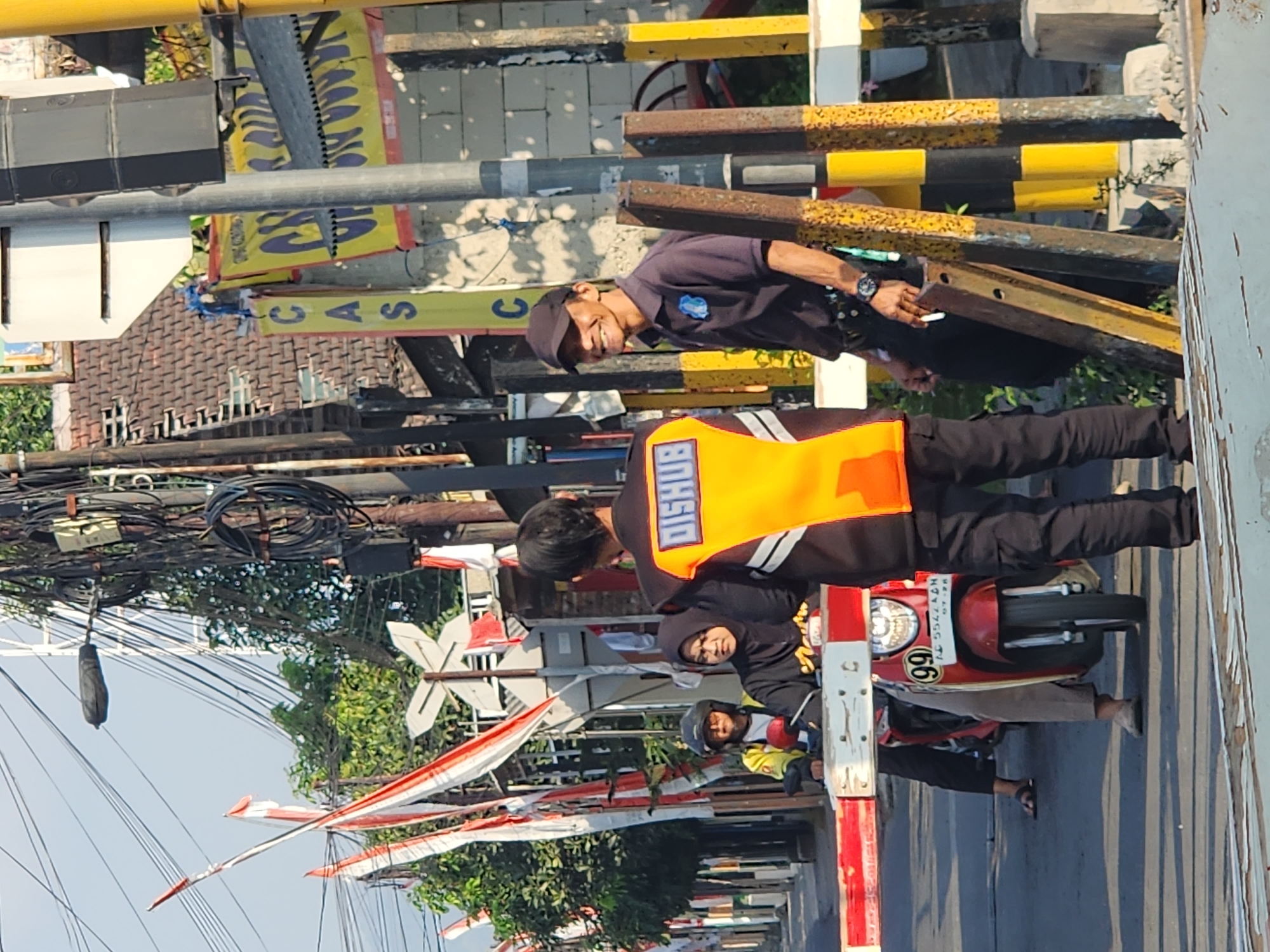 Two men, one in an orange vest with "DISHUB" written on the back and the other in a dark blue polo shirt and black cap, stand near a railway crossing in Kediri City, Indonesia. The man in the orange vest is facing away from the camera, while the man in the blue shirt is facing left, smiling and holding a cigarette. In the background, two individuals on a red scooter are waiting at the crossing, indicated by the lowered red and white barrier. The environment appears to be an urban street with overhead electrical wires, trees, and buildings. The overall scene captures a moment at a functioning railway crossing with people observing or waiting.