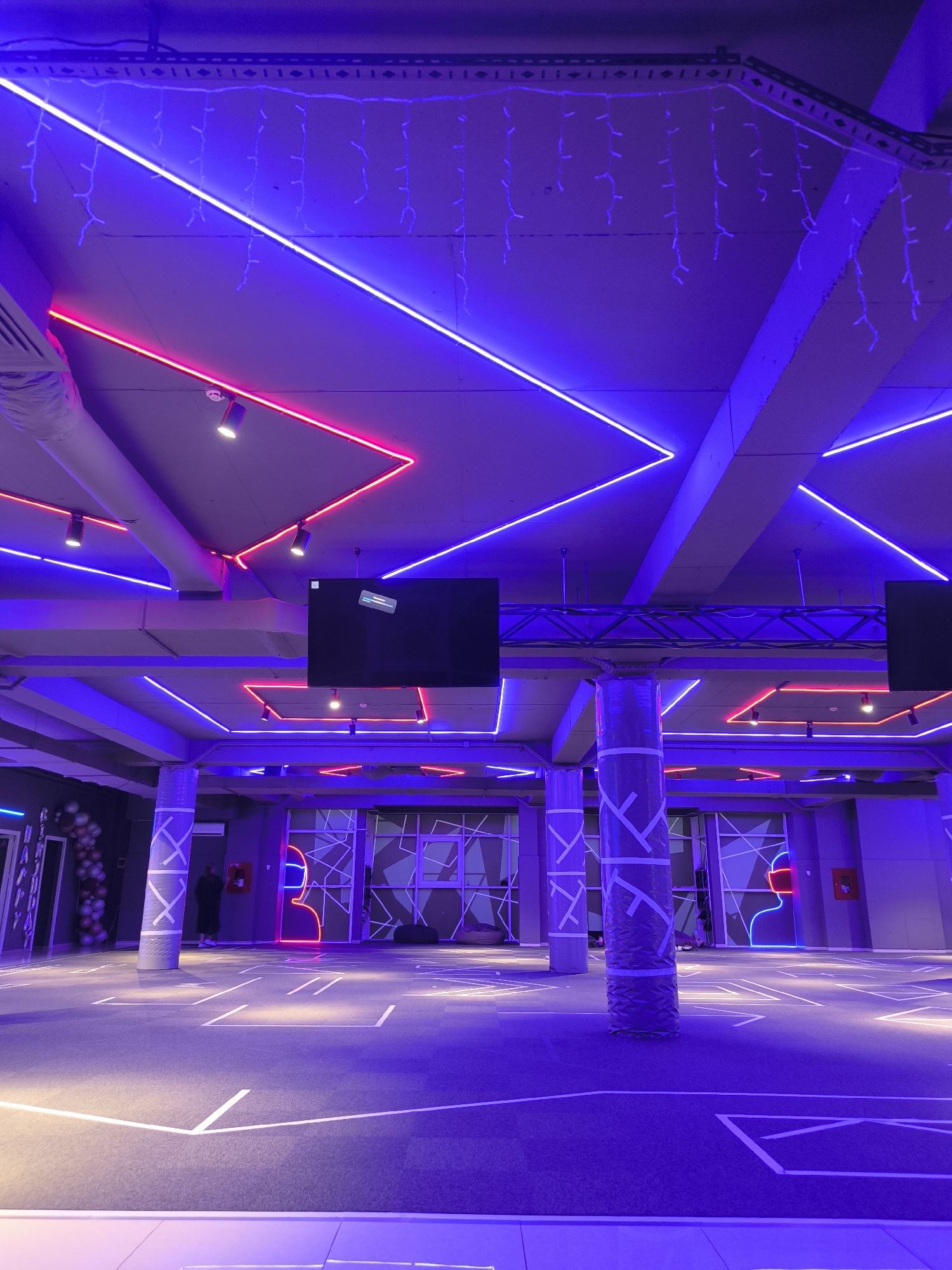 The image depicts a modern indoor space illuminated by vibrant neon lights in shades of blue and pink. The ceiling is adorned with diagonal lines of blue neon and some red neon accents, along with string lights that appear to be icicle lights. Several spotlights are also visible on the ceiling. Two large black screens are mounted on the ceiling structure. The walls feature geometric patterns, and there are cylindrical pillars wrapped in a reflective material with a geometric pattern. The floor is dark and marked with white lines, suggesting a designated area or game space. In the background, a person is visible near a red fire extinguisher mounted on the wall. To the left, there's an archway with balloons, possibly indicating an entrance or decoration for an event. The overall ambiance is futuristic and energetic. While the exact location in Chișinău, Moldova, is not specified, the scene suggests a recreational or entertainment venue, possibly a laser tag arena, escape room, or similar interactive experience center.