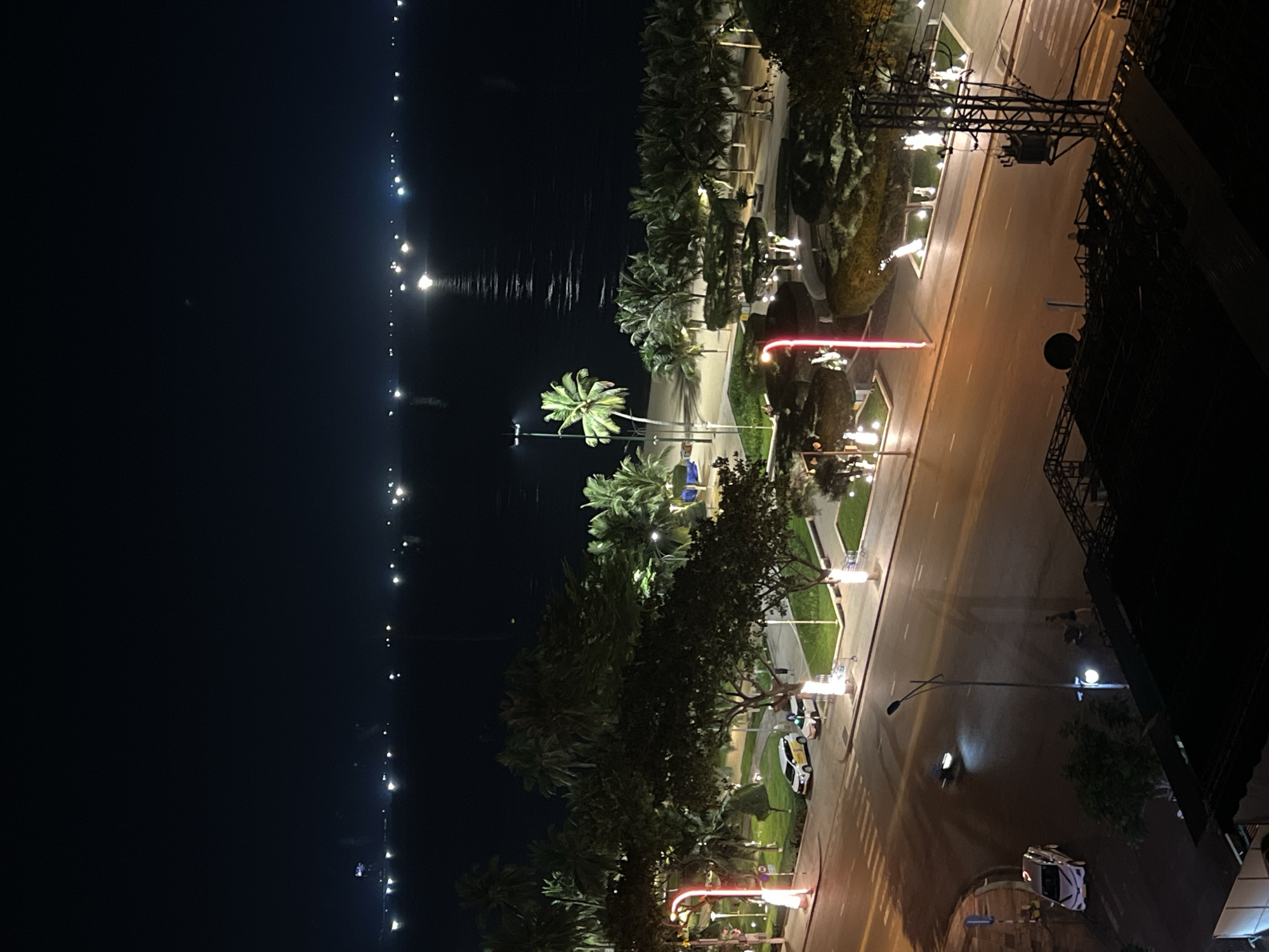 This image captures a nighttime view of a coastal promenade in Nha Trang, Vietnam. The scene is illuminated by artificial lights from street lamps, decorative lighting in parks, and distant city lights reflecting on the calm sea. The foreground features a paved road with a pedestrian crossing, where a white car and a motorcycle are visible. Beyond the road, a well-maintained park with manicured hedges and palm trees lines the promenade. Several pedestrians are seen walking along the illuminated paths. The sea stretches out to the horizon, dotted with distant lights that suggest a bustling cityscape or harbor. The overall atmosphere is tranquil and vibrant, showcasing Nha Trang's nocturnal beauty.